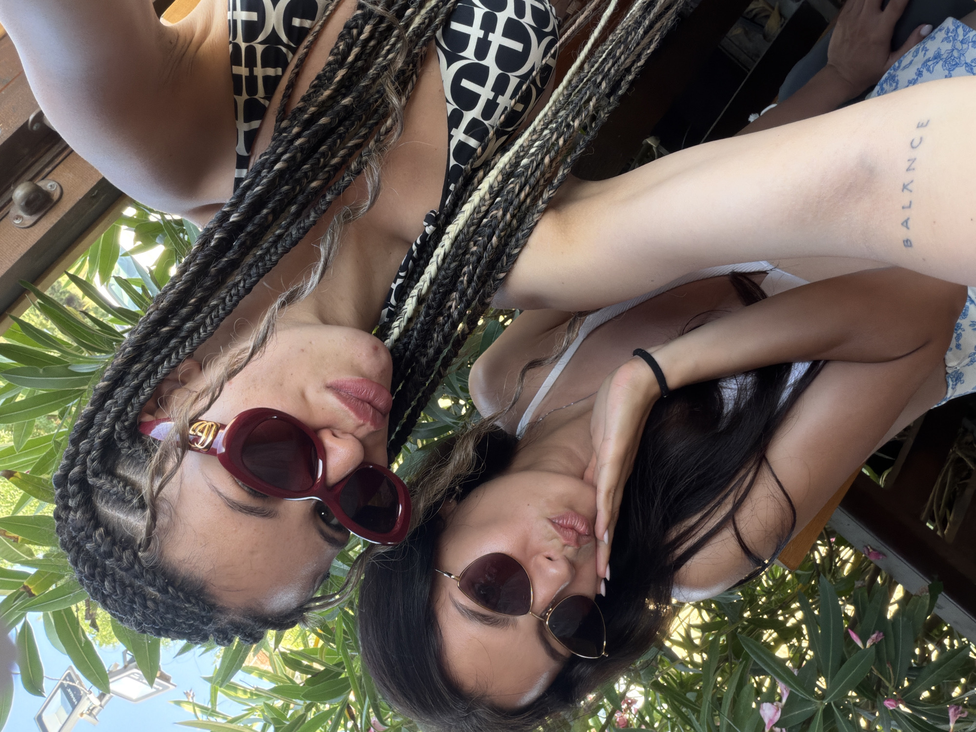 Two young women are taking a selfie outdoors. The woman on the left has dark hair, is wearing sunglasses, and a white bikini top. She is puckering her lips and resting her cheek on her hand. The woman on the right has braided hair with blonde highlights, is wearing large, dark red sunglasses, and a black and white patterned bikini top. She is also puckering her lips. They are positioned in front of lush green foliage and a wooden structure, suggesting an outdoor recreational area, potentially near a beach or resort. The lighting indicates it is daytime.