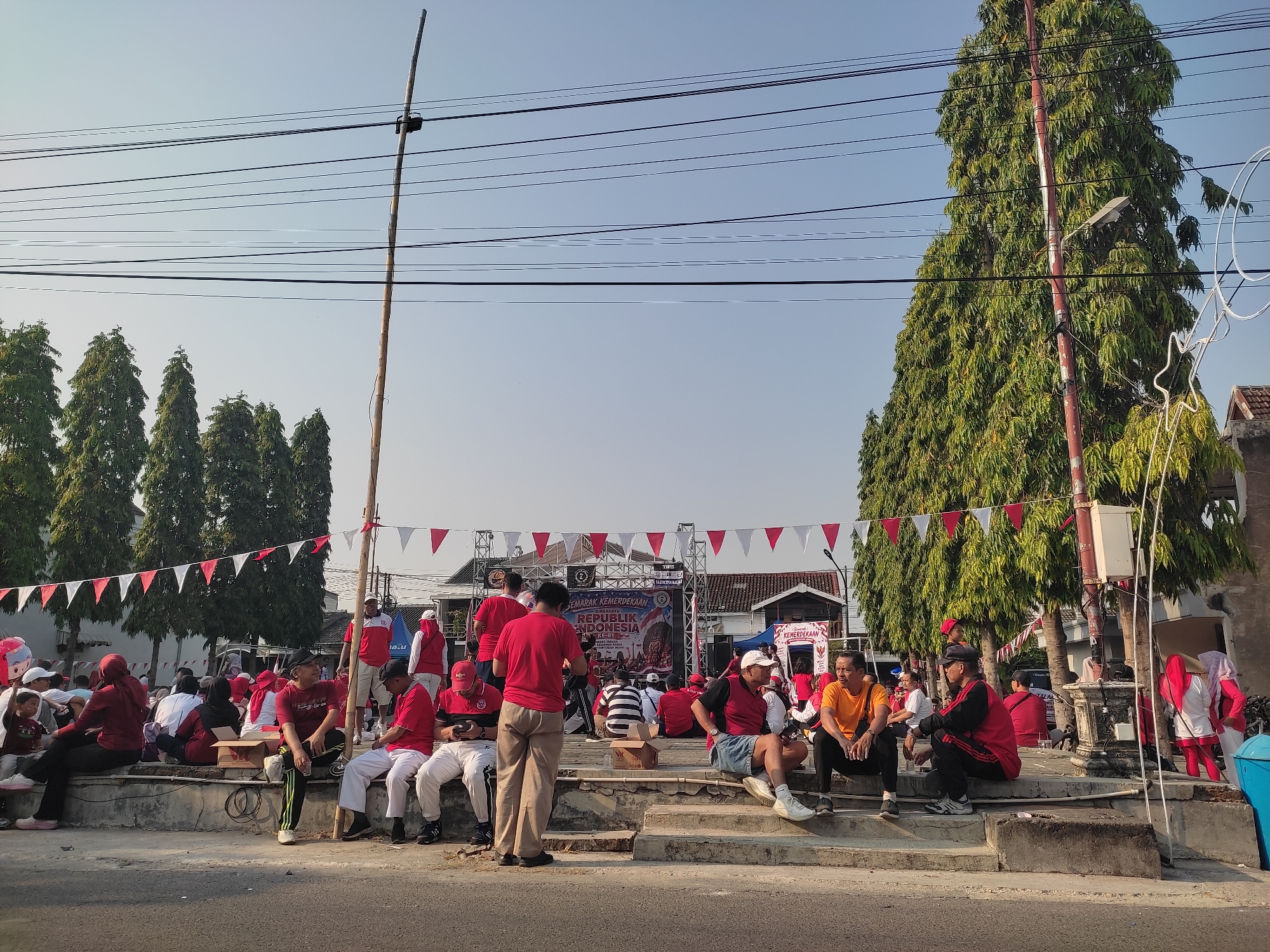 This image depicts an outdoor gathering in Ponorogo, Indonesia, likely celebrating Indonesian Independence Day, as evidenced by the red and white decorations and banners with the text "DIRGAHAYU REPUBLIK INDONESIA." The scene is set on a street with a stage in the background and a large number of people, mostly dressed in red and white attire, seated on concrete steps. Trees line the background, and utility poles with numerous wires are visible against a clear sky.
In the foreground, several individuals are seated on the steps. On the far left, a group is gathered, some appearing to be watching the stage. Moving towards the center, men in red shirts and white pants are seated, some engaged with their phones. One man in a red shirt and khaki pants stands in the center, facing away from the camera. Further to the right, more people are seated on the steps, including a man in an orange t-shirt and black pants, and another in a red and black jacket.
The stage area features large speakers, lighting equipment, and promotional banners. Red and white triangular flags are strung across the scene, adding to the festive atmosphere. The overall impression is a public event with a relaxed, community-oriented feel, likely a local celebration or performance. The presence of utility poles and the architectural style of the buildings in the background suggest an urban or semi-urban environment within Ponorogo.