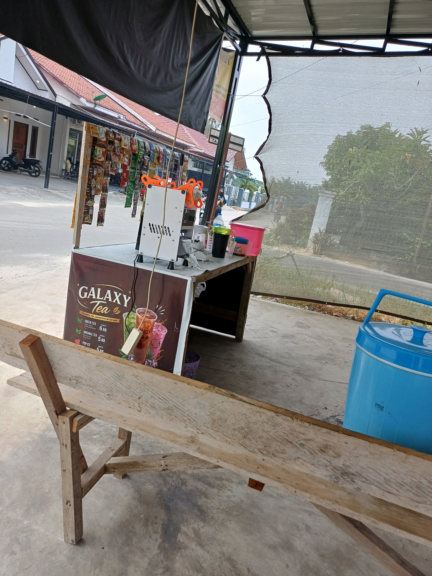 The image depicts an outdoor beverage stand, likely located in Dumai City, Indonesia, based on context. In the foreground, a rough-hewn wooden bench is positioned on a concrete surface. Behind the bench, a vendor's stand is set up under a black canopy. The stand features a white sealing machine for cups, and a banner advertising "GALAXY Tea" with different flavor options like "GREEN TEA," "ORIGINAL TEA," and "POP ICE." Various packaged snacks are hanging from a rack above the counter. To the right of the stand, a bright blue insulated cooler is visible. The background shows a street with buildings and some vegetation, suggesting a commercial or residential area. The overall scene is informal and appears to be a typical street-side stall.