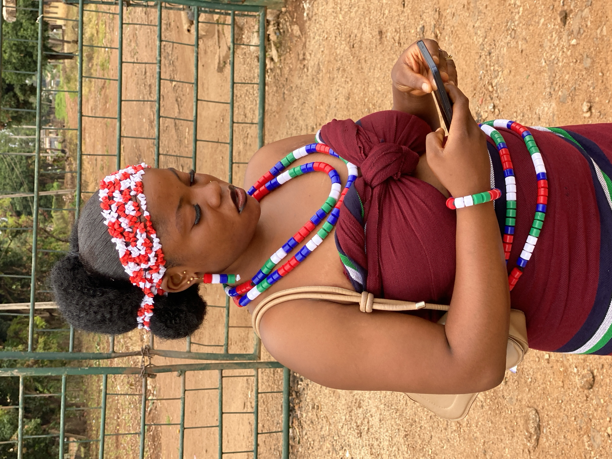 A young Black woman stands outdoors, presumably in the city of Maigizo, Nigeria, operating a black cell phone in her right hand. She is wearing a maroon top and a beaded headband with alternating red and white beads. She also has a beaded necklace and bracelet with red, white, and green beads. Her hair is styled in a bun. She is wearing a tan purse, and is in front of a green metal fence. The ground is dirt, and there are trees in the background.