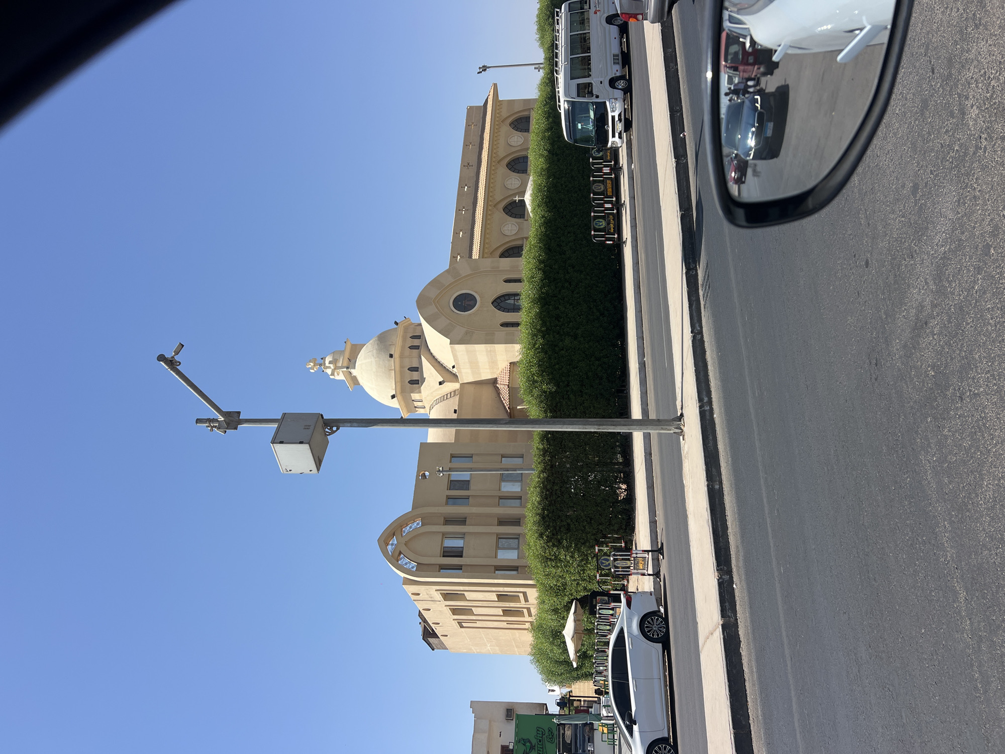 This is a photograph taken on a sunny day in the city of Sharm El Sheikh, Egypt, specifically depicting a view of the Coptic Orthodox Cathedral of the Archangel Michael. The Cathedral is a large, sand-colored building with a prominent dome and arched windows. It is surrounded by a dense, green hedge. In the foreground, a white sedan is parked on the side of the road, and a white minibus is driving past. The road in front of the Cathedral appears to be a main thoroughfare with other vehicles visible in the distance. A lamppost with a security camera is also present. The scene is viewed from the perspective of a car, with the side-view mirror reflecting other vehicles on the road, including a dark sedan and what appear to be several pedestrians. The sky is clear and blue.