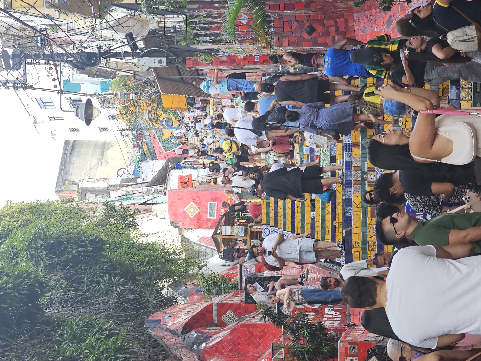 The image shows a view of the Escadaria Selarón (Selarón Steps) in the Lapa neighborhood of Rio de Janeiro, Brazil. This landmark is composed of colorful tiles, ceramics, and mirrors. A large group of people are walking or standing on the steps, many seemingly tourists, dressed in casual clothing. The steps appear crowded with people of varying ethnicities and ages.
The steps are decorated with tiles of different colors and patterns, including national flags and other designs. Surrounding the steps are buildings, trees, and overhead utility wires and cables. The sky is overcast, and the weather appears to be daytime. The foreground and background are moderately blurred, giving the focus to the people and the steps.