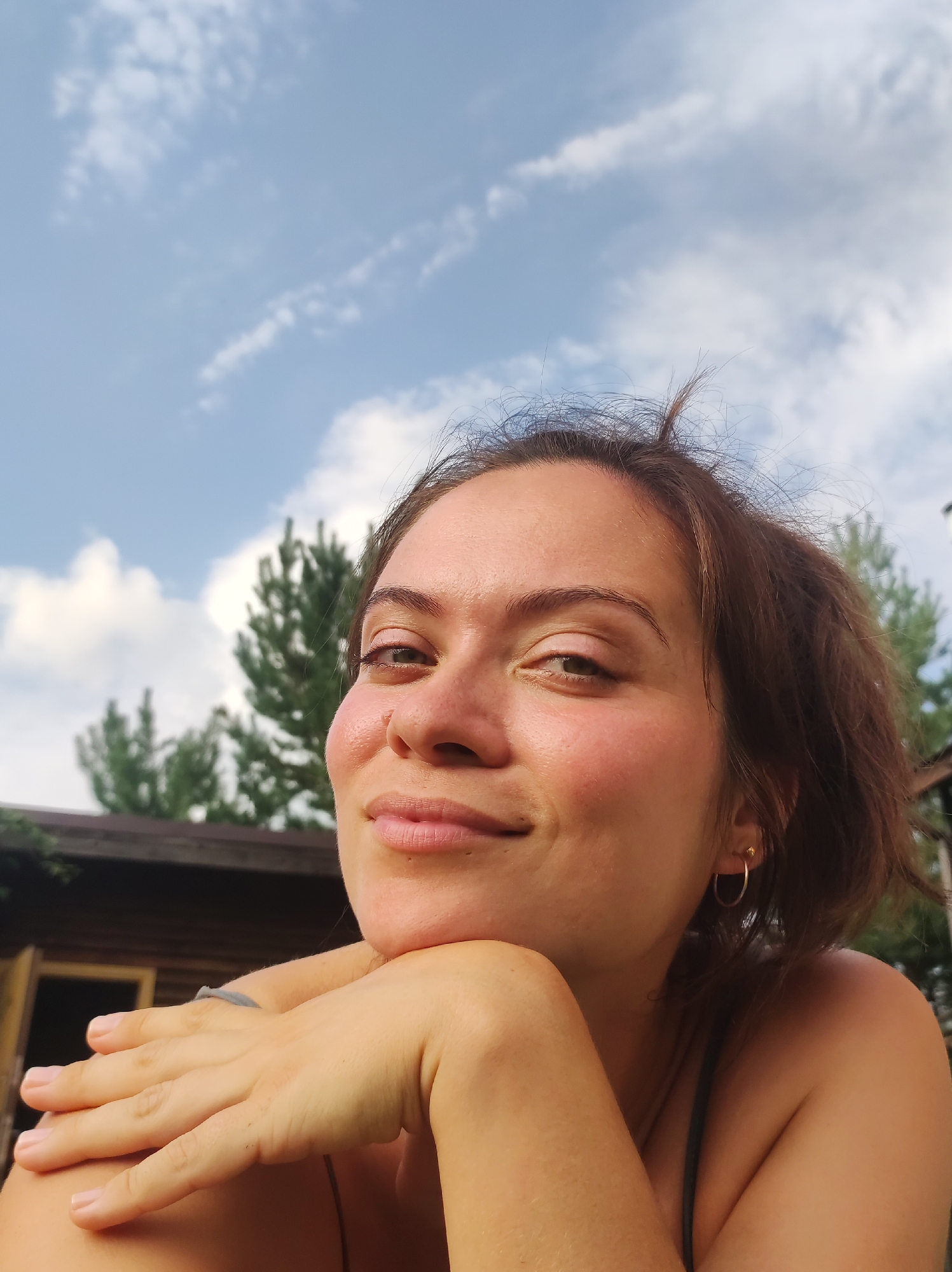 A young woman is pictured in a close-up shot, with her face angled slightly towards the camera. She is outdoors, with a blue sky and scattered clouds visible behind her, along with trees and a wooden structure that appears to be a small house or cabin. The woman has brown hair, which is slightly messy and windswept, and she is wearing a subtle hoop earring. Her hand is resting on her chin, and she has a gentle smile on her face. The lighting suggests it is daytime, possibly late afternoon, with the sun casting a warm glow on her skin. The scene evokes a relaxed, natural atmosphere, possibly from a rural setting or a vacation spot. The location is identified as Фоки, Russia.
