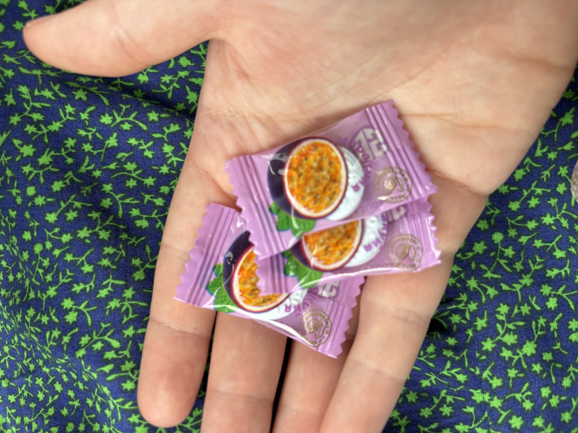 A close-up overhead shot shows a person's open palm resting on a navy blue fabric with a green floral pattern. Three small, individually wrapped purple candies, each featuring an image of passion fruit, are held within the palm. The candies are slightly overlapped, with the front two being more visible. The fabric background is slightly out of focus.
