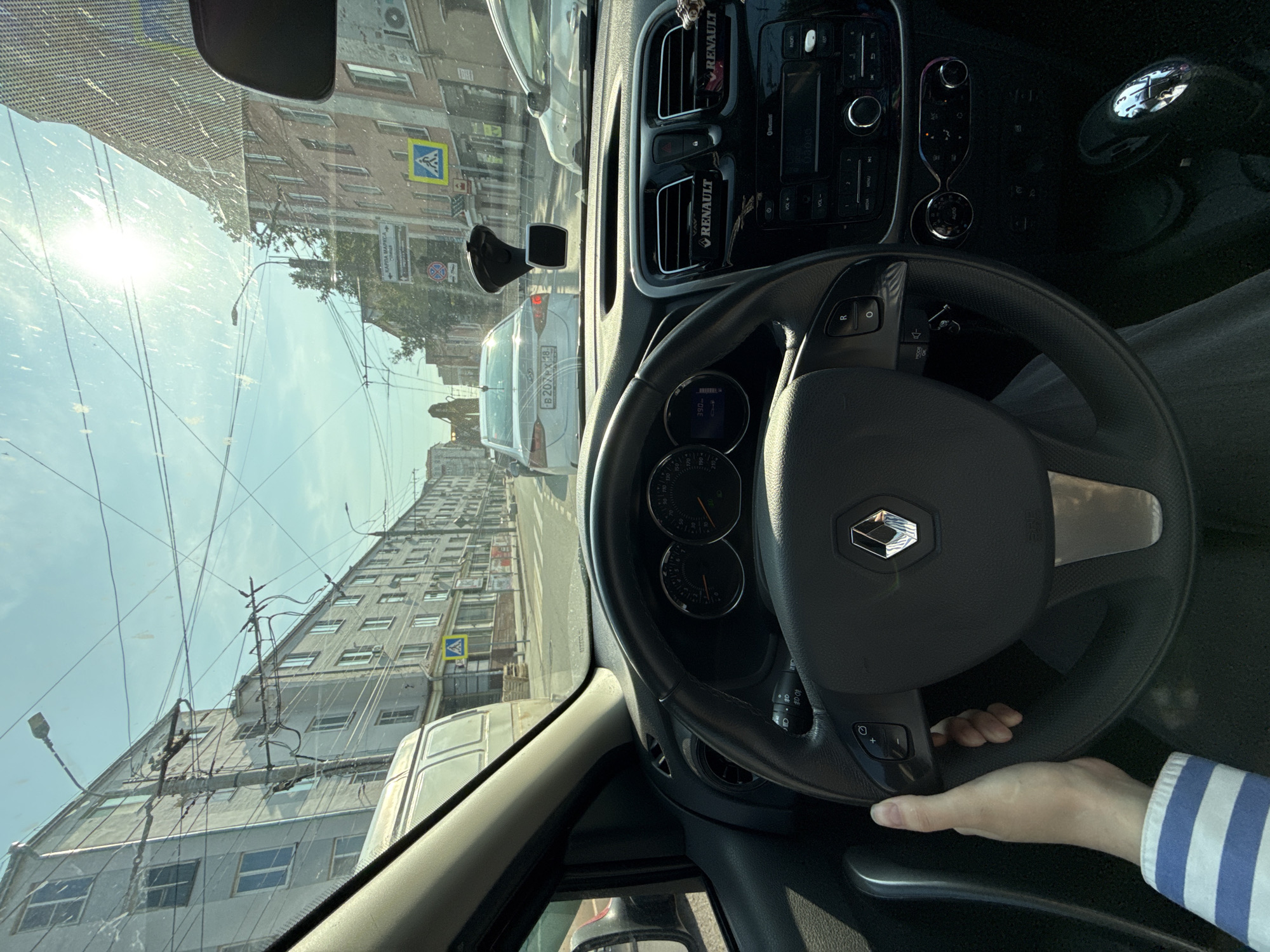The image is a first-person perspective from inside a car, likely a Renault, driving on a street in Izhevsk, Russia, on a sunny day. The driver's hand is visible on the steering wheel, and the dashboard displays include the speedometer and tachometer. The car is in traffic, with other vehicles ahead, including a white sedan with a license plate that appears to be in Cyrillic script.
The surrounding environment consists of multi-story apartment buildings lining the street, characteristic of Soviet-era architecture, with many windows and balconies. Overhead, a complex network of wires, likely for trolleybuses or trams, crisscrosses the sky. Street signs are visible, including a pedestrian crossing sign. The sun is bright, causing some lens flare on the windshield. The overall impression is a typical urban street scene in a Russian city.