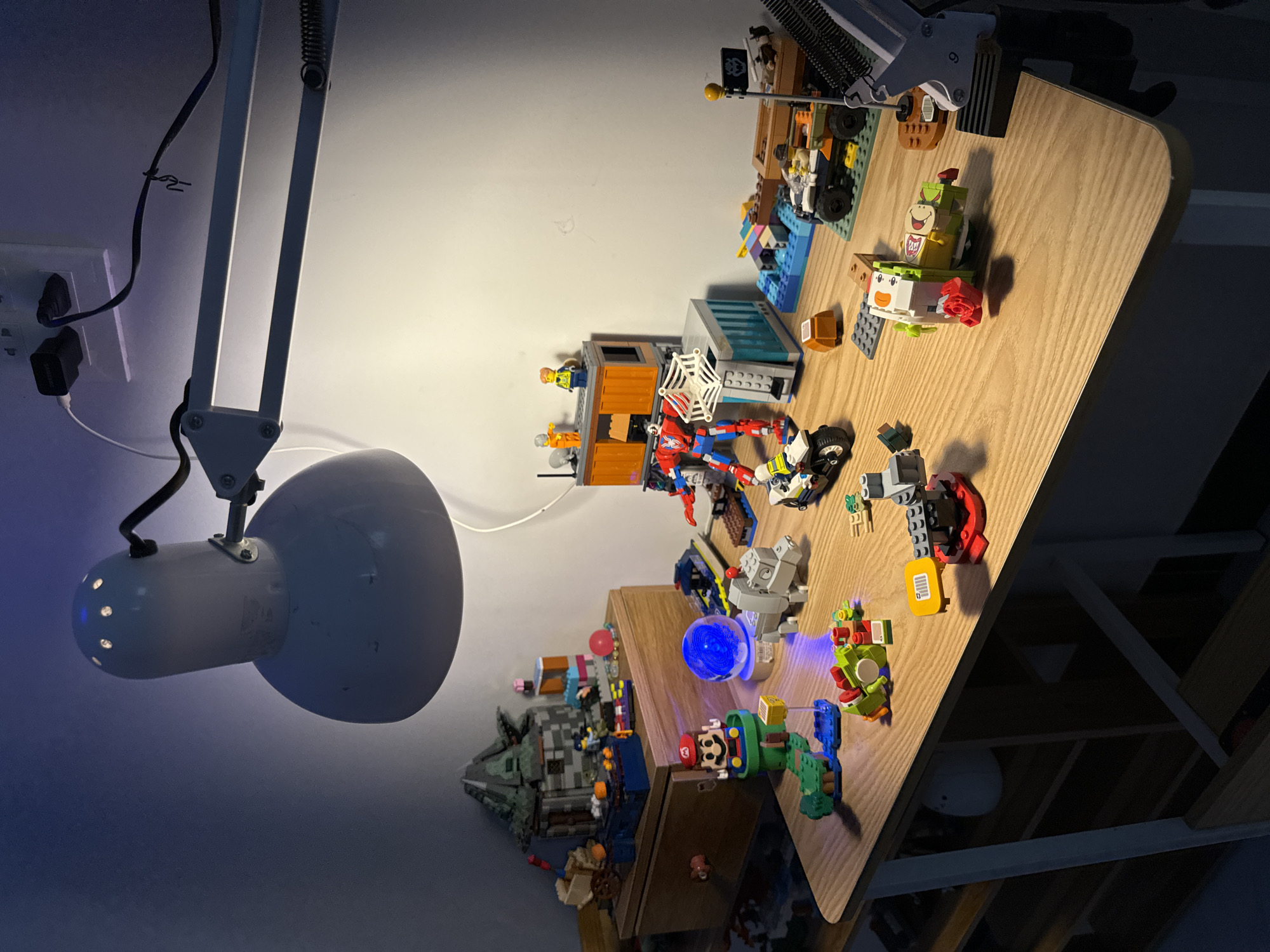 The scene depicts a desk covered with various Lego sets. A white articulated lamp is positioned on the left side, illuminating the desk and the surrounding wall. The desk surface is light brown and cluttered with Lego constructions, including buildings, vehicles, and figures. Some Lego sets are identifiable, such as those from Marvel and Super Mario. The background shows a light-colored wall and parts of the room, including a power outlet and a glimpse of shelves or a bookcase. The lighting in the room is a mix of natural and artificial, creating shadows and highlights on the objects. The scene is cluttered and appears to be a workspace or hobby area.
The location of Mũi Né, Vietnam, is not evident in the image.