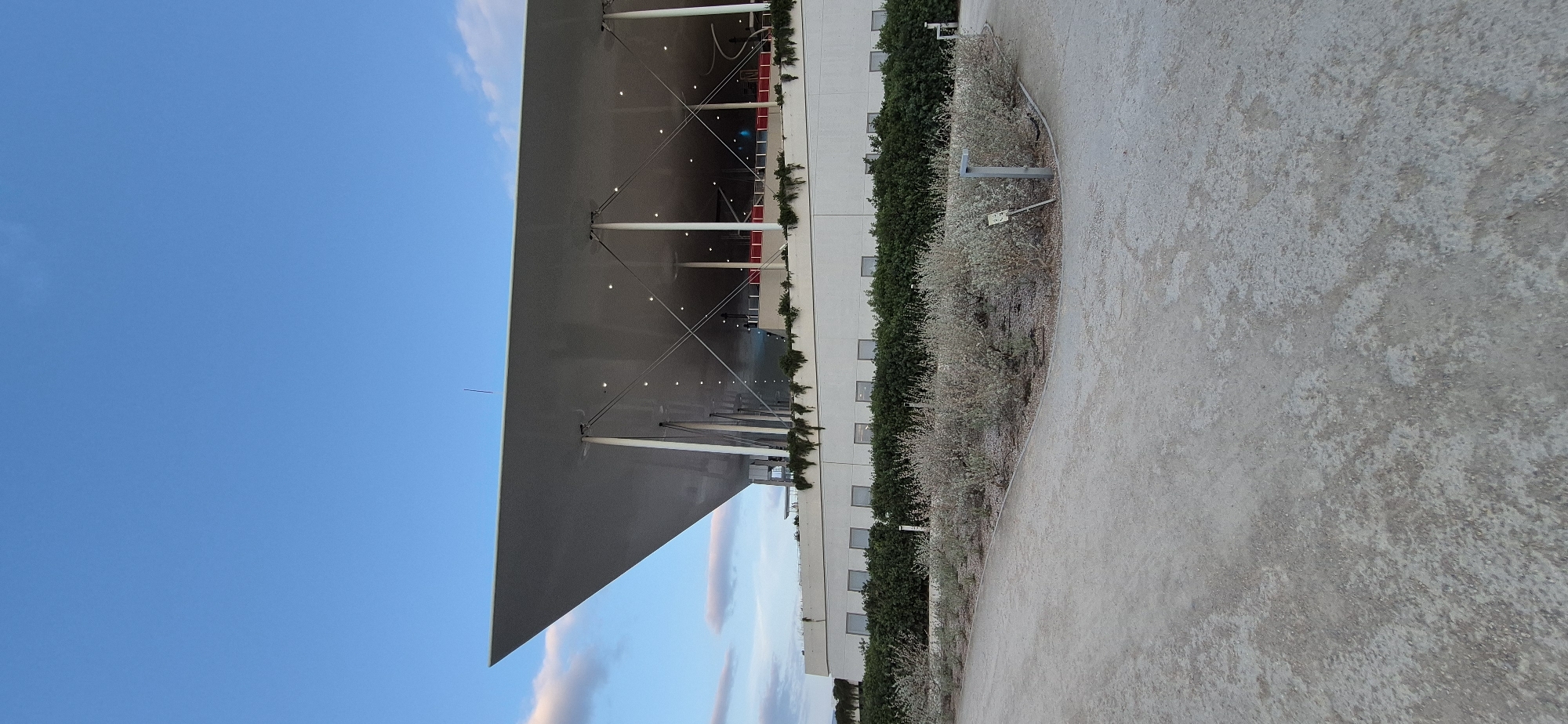 The image depicts the Stavros Niarchos Foundation Cultural Center (SNFCC) in Kallithea, Greece. The perspective is from a pathway in front of the building, which features a large, overhanging roof supported by columns. The building's facade is visible, with a row of windows and lush green bushes and lighter, dry shrubbery in the foreground. The sky above is clear blue with some wispy clouds. The overall impression is of a modern architectural complex set within an outdoor landscape.