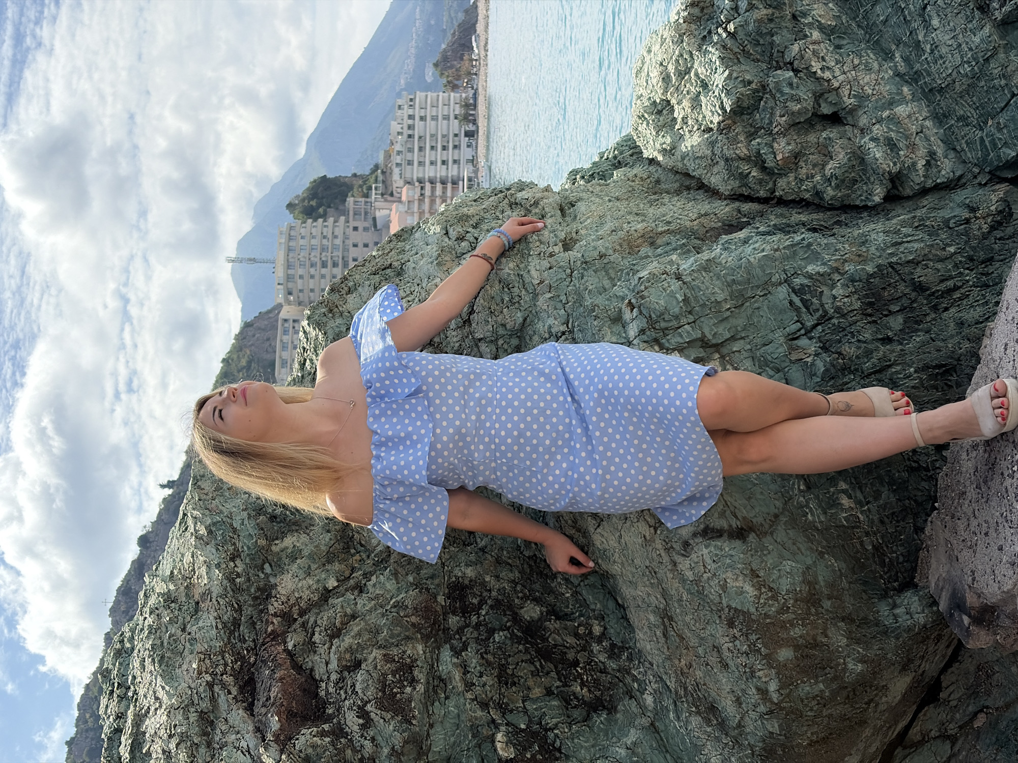 A woman wearing a blue and white polka-dot dress is lying on a large, craggy rock formation. She has blonde hair and is wearing light-colored sandals. In the background are buildings, a mountain range, and the sea. The location is Rafailovići, Montenegro.