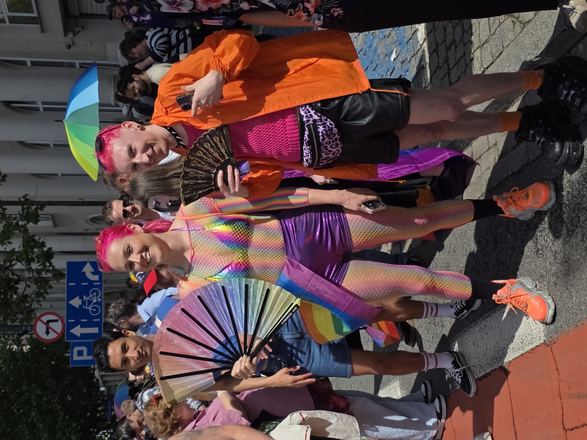 The image captures a vibrant scene during a Pride parade in Warsaw, Poland. In the foreground, two individuals are the primary focus. The person on the left, a woman with bright pink hair, wears a shimmering rainbow-patterned mesh top and metallic purple shorts. She is holding a large, colorful fan. To her right, a person also with bright pink hair, sports a hot pink knitted top and black leather shorts, with an orange windbreaker over their shoulders. They are holding a black fan in their left hand and a mobile phone in their right. A leopard-print fanny pack is worn around their waist.
Behind them, a crowd is visible, with many participants holding rainbow flags and fans, indicative of a Pride event. The backdrop features a street scene with buildings and trees, characteristic of an urban environment. Traffic signs are visible on a pole in the background. The ground is paved, with a pedestrian crossing marked by red and white lines. The overall atmosphere appears festive and celebratory.