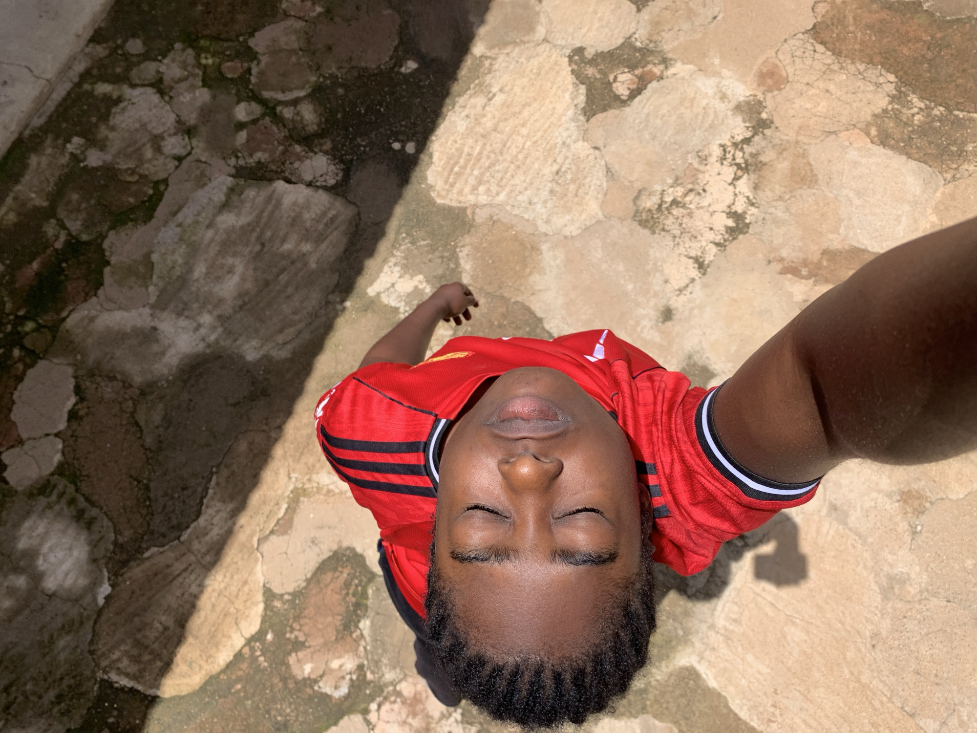 This is an upward-facing selfie taken in Rafin Par, Nigeria. The subject is a young person wearing a red t-shirt with black and white trim on the collar and sleeves. They have their eyes closed, and their head is tilted back, looking upwards. Their hair is styled in braids.
The setting appears to be an outdoor area with a ground made of rough, uneven flagstones. Some of the stones are light brown and beige, while others are darker and appear damp or mossy. A strong shadow from an unseen object falls diagonally across the ground and the subject, suggesting it is daytime with bright sunlight. The overall mood of the image is peaceful, with the closed eyes and upward gaze conveying a sense of reflection or relaxation. No visible text is present in the image.