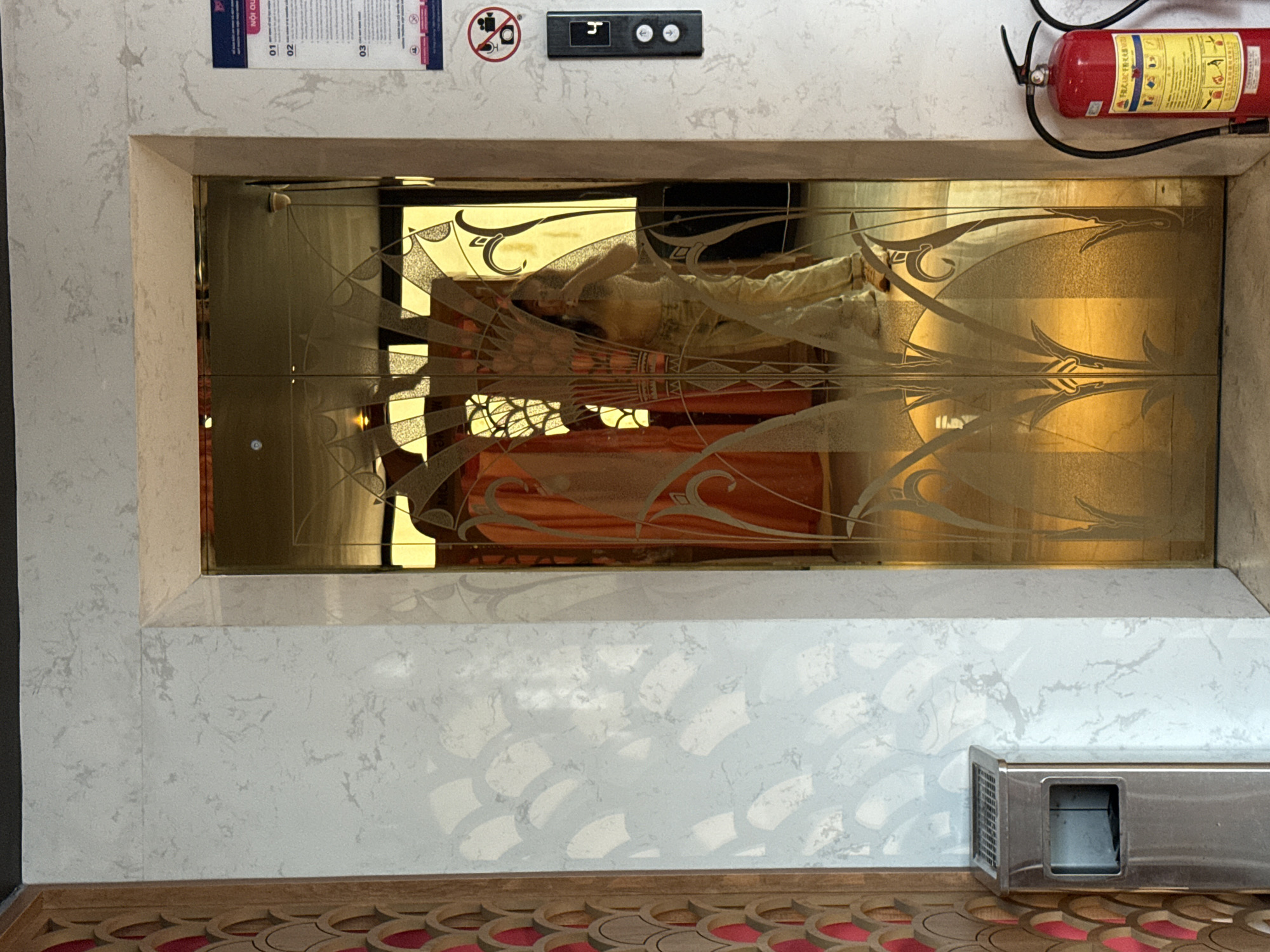 A mirrored and ornate elevator door is set into a wall patterned with gray and white faux marble. Above the door are a fire extinguisher and a small panel with elevator controls and signage. Reflected in the elevator door is a person wearing a white outfit lying on an orange surface. Below the door, there is a gray metal object and patterned flooring with red accents. The image was taken in Bac Nha Trang Ward, Vietnam.