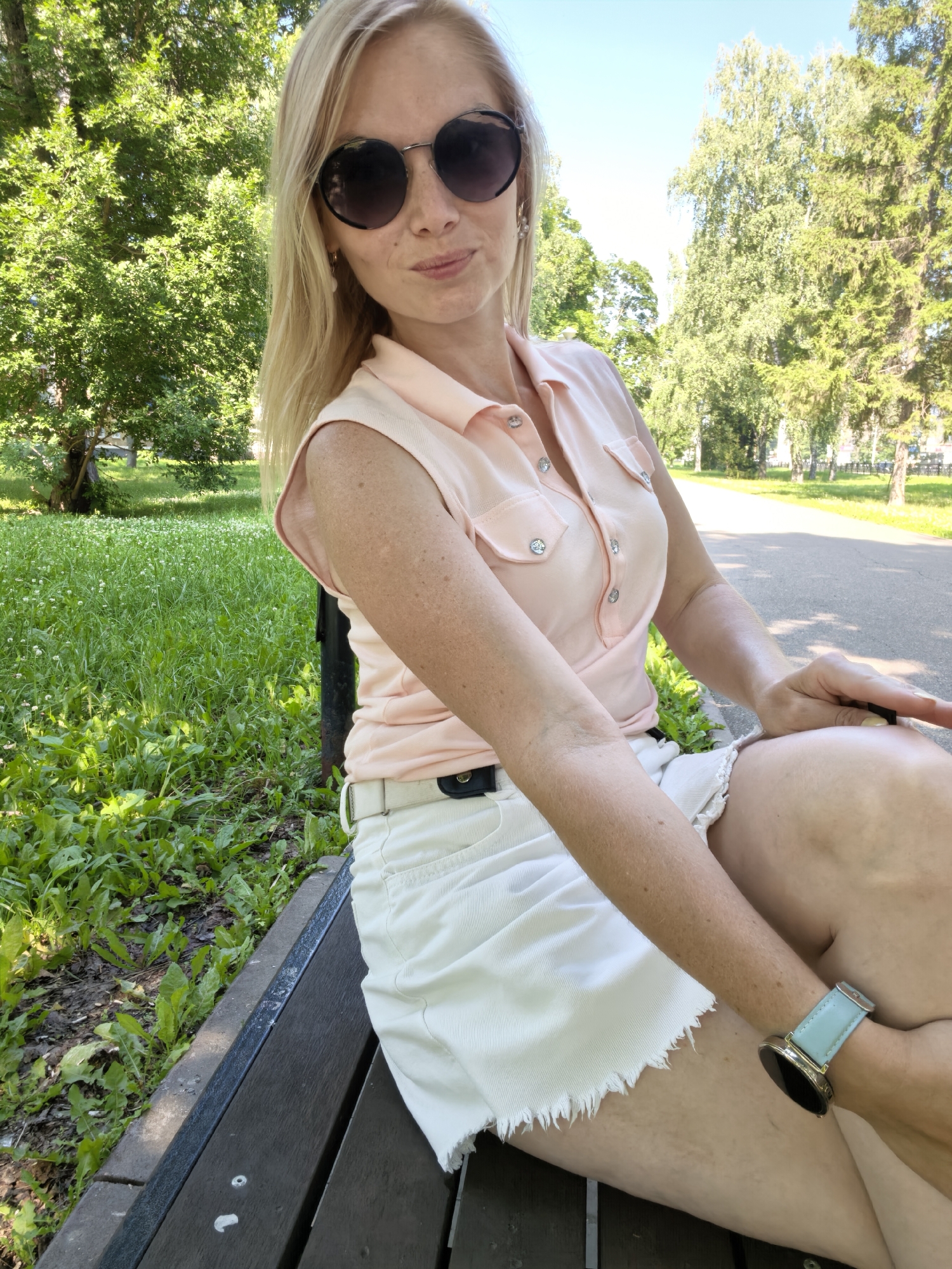 A blonde woman wearing sunglasses, a pink sleeveless shirt, and a white skirt sits on a park bench. She is outdoors in a park with trees and grass visible in the background. The image appears to be taken during the daytime.