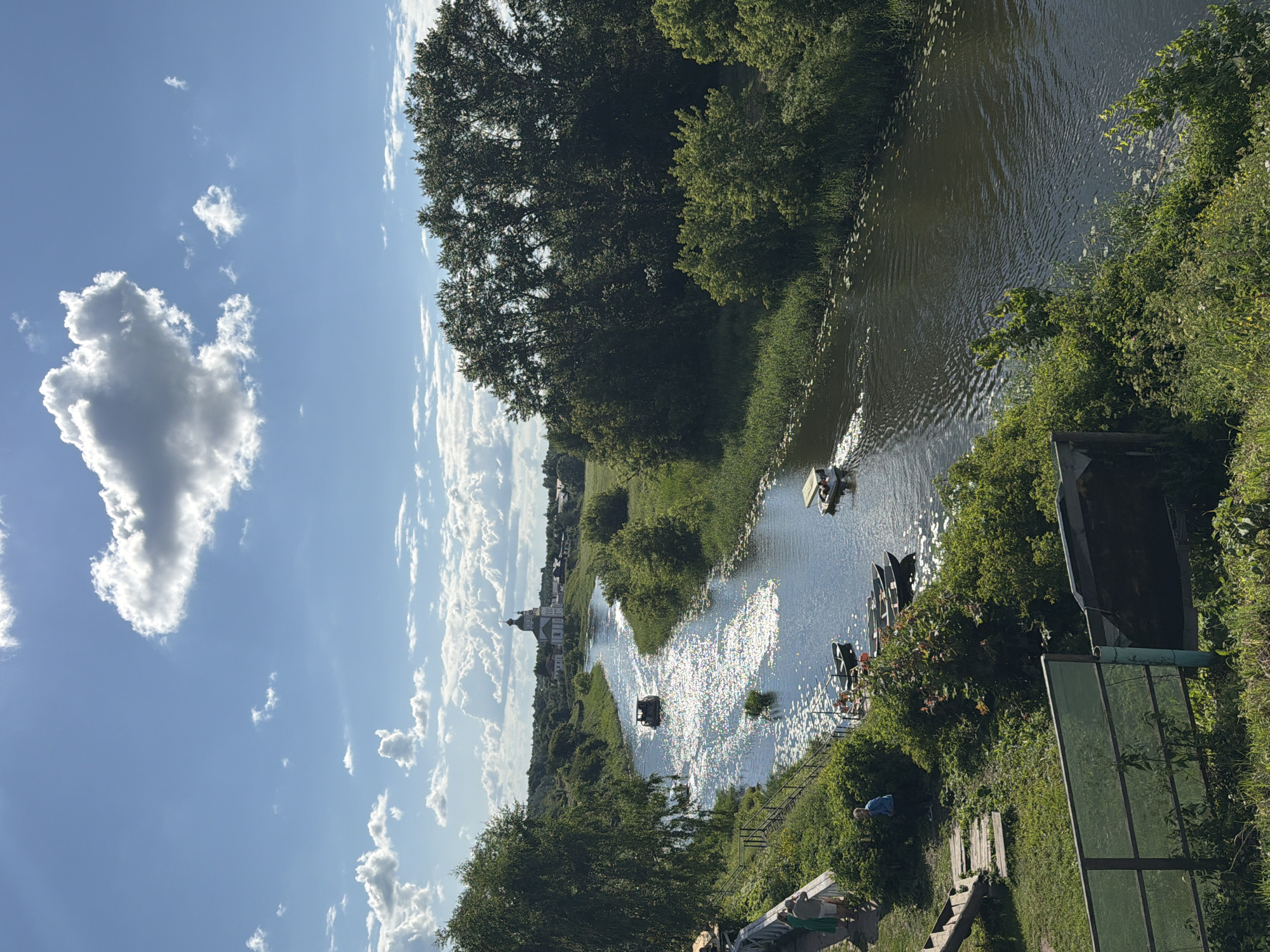The image captures a scenic view of the Kamenka River flowing through the city of Suzdal, Russia, on a bright, sunny day. The river is wide and reflects the sunlight, creating a shimmering effect on the water's surface. Several boats are visible on the river, including a larger tour boat and smaller rowboats. The riverbanks are lush and green, with dense vegetation and trees lining both sides. In the distance, a prominent white church with a green dome stands out against the blue sky, adding a touch of architectural beauty to the natural landscape. The sky is a vibrant blue, dotted with fluffy white clouds. On the left bank of the river, there is a wooden walkway and steps leading down to the water, where several people are gathered. One person is standing near the water's edge, looking out at the river. Another group of people are walking along the walkway. The overall impression is one of a peaceful and picturesque summer day in a historic Russian city.