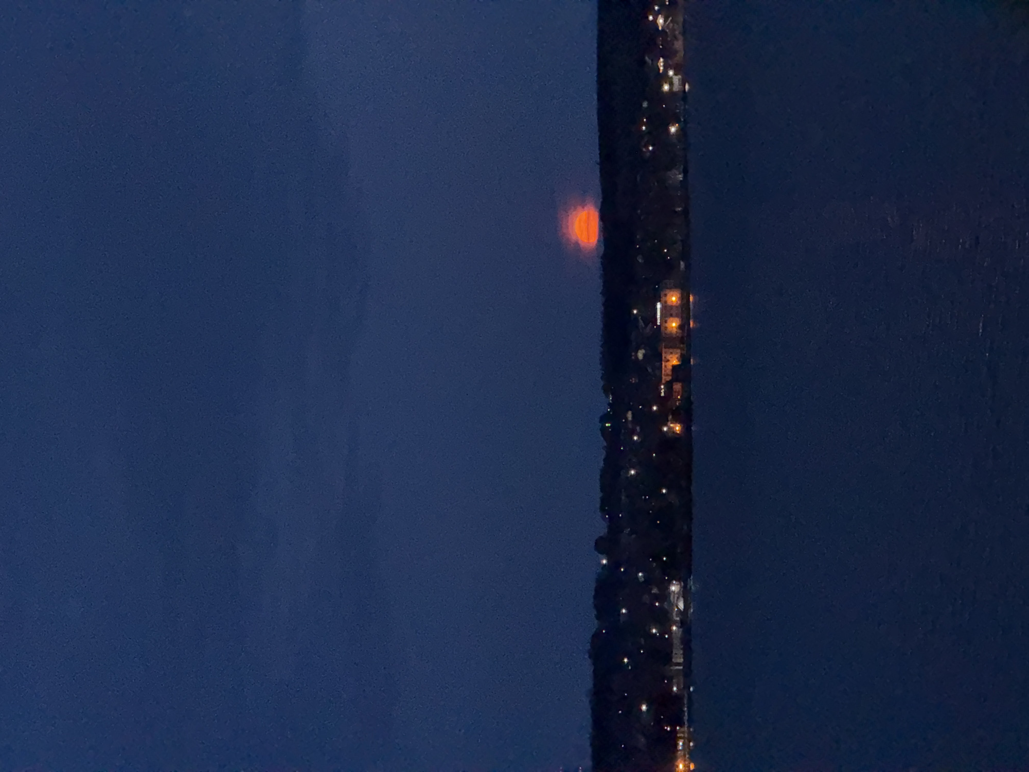 The image shows a nighttime view of Votkinsk, Russia. A section of city buildings with visible lights occupies the right side of the frame. To the left of the buildings, the night sky is dark blue. In the center, a faintly visible orange or red circular object that might be the moon or a light source behind atmospheric haze.