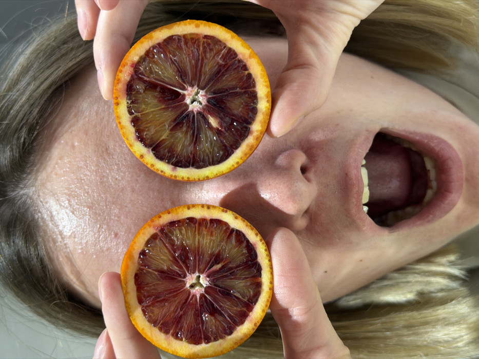 A close-up, portrait-oriented image depicts a person holding two halves of a blood orange over their eyes. The individual's light-colored hair is visible at the top and sides of the frame. Each orange half, showcasing a vibrant radial pattern of dark red and orange flesh surrounded by a lighter orange rind, is held in place by the subject's fingers. Below the oranges, the person's nose is visible, and their mouth is wide open in a distinct "O" shape, revealing upper and lower teeth, the tongue, and the back of the throat. The expression conveyed is one of surprise or playful astonishment. The background is indistinct and uniformly light-toned, suggesting an indoor or studio setting. The specific location within the city of Chaikovsky, Russia, cannot be visually determined from the details present in this image.