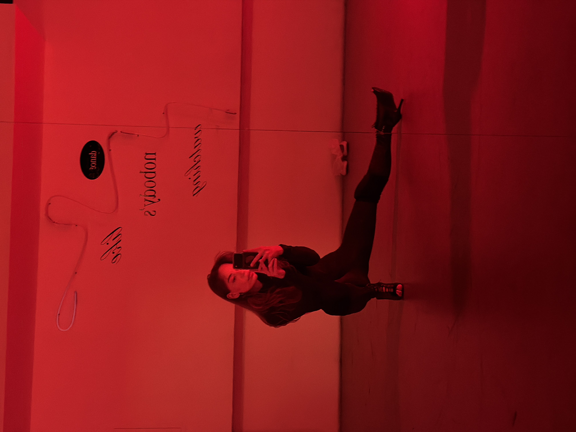 A single female subject is observed taking a mirror selfie in a room illuminated with monochromatic red lighting. The subject is positioned in a low squat or lunge, with her left leg bent and her right leg extended horizontally to the right. She is wearing a form-fitting black long-sleeved top, black leggings, and black open-toed high-heeled shoes with ankle straps. Her long, dark hair falls over her right shoulder. She holds a dark-colored smartphone in both hands in front of her face, appearing to look at the screen.
The background wall is a large reflective surface, likely a mirror, displaying reversed text from neon signs. Prominently visible are the words "dance" (within a black oval), "nobody's," and "painkiller." A pair of light-colored athletic shoes are visible on the floor against the wall in the upper right background. The floor is a dark, smooth surface. This environment is identified as the Nobody's Painkiller Dance Studio, located at Presnenskaya Naberezhnaya 10, Block 2, Moscow, Russia.