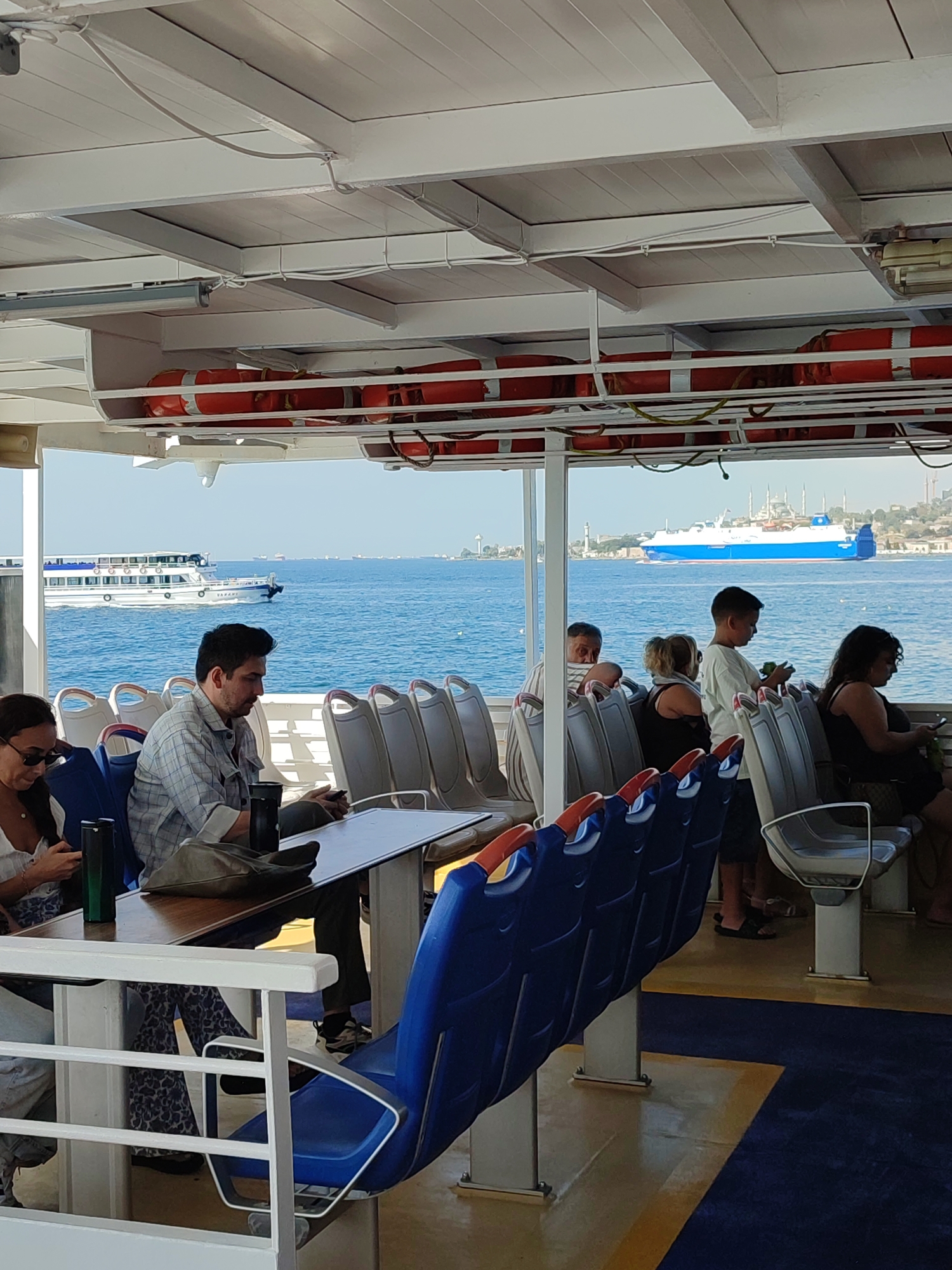 Onboard a ferry, several passengers are seated and standing, looking out at the sea. In the foreground, a man in a checkered shirt sits at a table, looking down. To his left, a woman in sunglasses is looking at her phone. In the background, a large blue and white ferry sails across the water, with the distinctive skyline of Istanbul, including mosques and minarets, visible on the horizon. The ferry interior features rows of blue and gray seats, some with orange accents, and overhead life preservers. The overall scene depicts a calm day at sea with passengers enjoying the view of the cityscape of Üsküdar, Turkey.