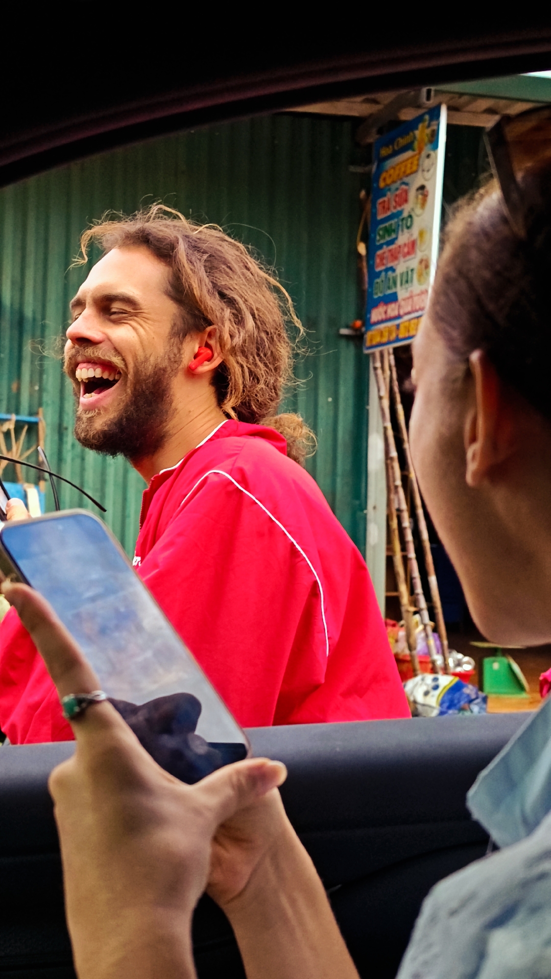 The image captures a candid moment in Xín Vàng, Vietnam, seen from inside a vehicle looking outwards. In the foreground, positioned in the lower left, a person's hands, wearing a silver ring with a green stone on the left ring finger, hold a dark-colored smartphone horizontally. The phone screen reflects what appears to be a cloudy sky or a distant outdoor scene.
To the left, a man with light brown, curly, shoulder-length hair tied back in a ponytail, and a beard, is laughing boisterously with his eyes closed and mouth wide open, revealing his teeth. He wears a bright red jacket with white piping details and has a red wireless earbud in his left ear. He appears to be holding a pair of glasses in his left hand, partially obscured by the phone in the foreground.
Behind the man, a green corrugated metal wall serves as a backdrop. To the right of the laughing man, a vertical white sign with blue Vietnamese text is visible, listing "COFFEE," "TRÀ SỮA" (Milk Tea), "SINH TỐ" (Smoothie), and "BIA" (Beer), indicating a local street vendor or establishment. Below the sign, several vertical poles and an assortment of items, including colorful bags and containers, are visible at ground level.
On the right side of the frame, partially obscured and seen from behind, is the head and shoulder of a second person, likely female, with brown hair tied back. Her right ear is visible, and she is wearing a light blue top. She is looking towards the laughing man, creating an implied interaction between the two individuals. The dark gray interior of a car door or window frame is visible along the bottom and right edge of the image.