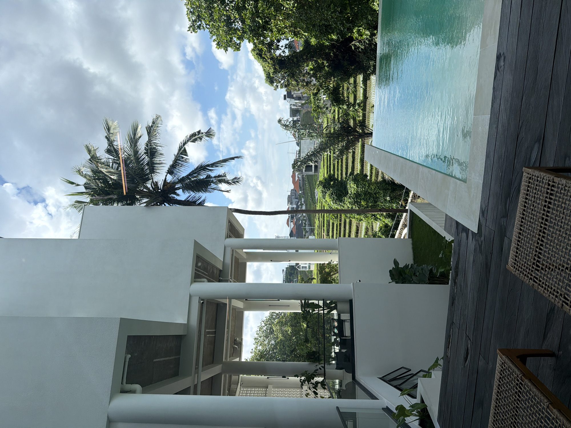 Captured in Mengwi, Indonesia, the image showcases an outdoor space featuring a contemporary architectural design. Predominantly white structures, including columns and walls, define the building's aesthetic. The scene includes a swimming pool with clear water, bordered by dark-toned wooden decking. The surroundings offer a view of lush green rice terraces, dotted with trees and residential buildings. A palm tree is visible against a backdrop of a blue sky with scattered clouds. Wicker chairs are partially visible in the foreground, set on the dark decking.