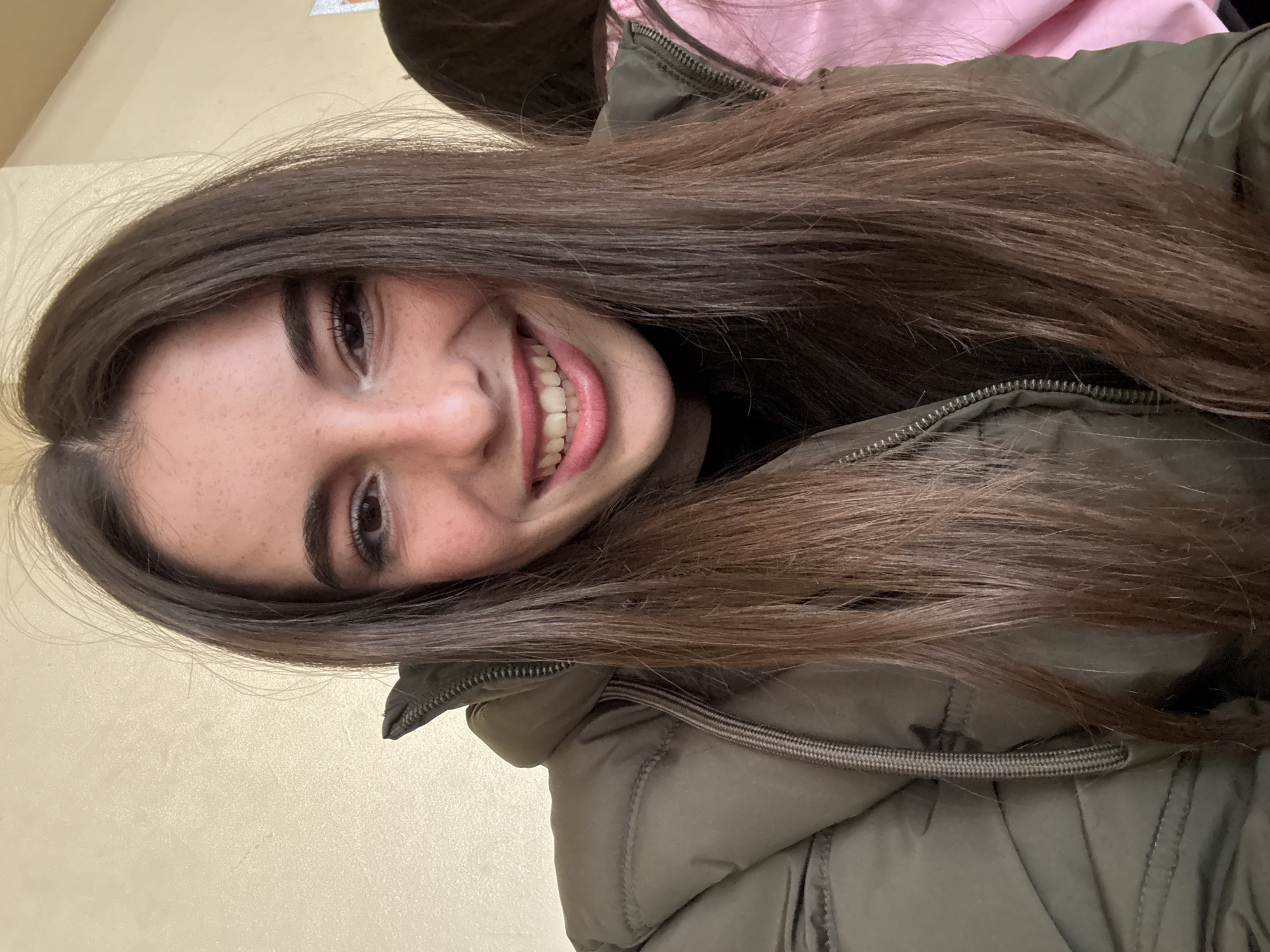 A woman with long brown hair is smiling at the camera. She is wearing a green puffy coat. The background is a light yellow wall. The image does not contain enough information to determine the location in Plovdiv, Bulgaria.