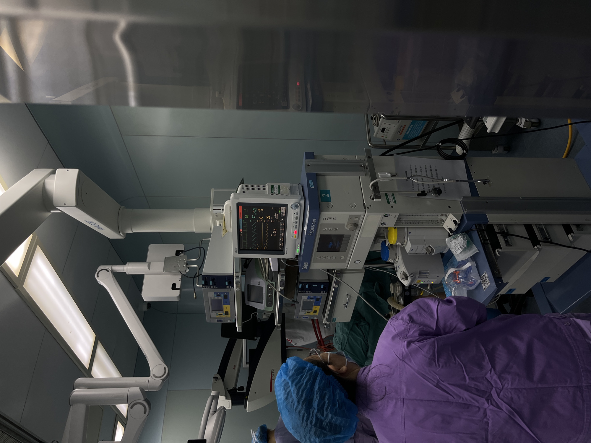 The image depicts a scene inside what appears to be a medical operating room. Dominating the view are various pieces of medical equipment, including monitors displaying vital signs, anesthesia machines (likely Dräger Fabius plus), and surgical lights mounted on articulated arms (possibly Leica F50). A medical professional wearing a blue surgical cap and purple gown is visible near the bottom of the image. The environment appears clean, with pale blue walls and stainless-steel surfaces. Some of the equipment has indicators labeled "EQUATOR".
There is nothing in the image that explicitly states that the location is in the city of Yuzhong District, China.