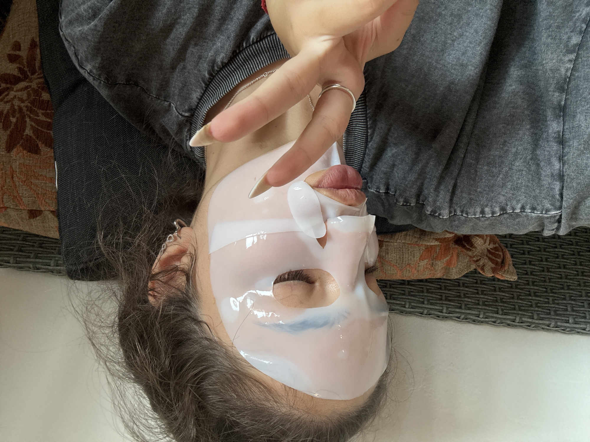 A young woman with her eyes closed and a white sheet mask on her face is shown lying on a couch. She is wearing a grey t-shirt, a silver ring on her left index finger, and a necklace. Her right hand is held up in front of her face, with her index and middle fingers extended in a peace sign. She has long, dark, messy hair. The couch is upholstered in a patterned fabric of orange and brown. The background is a plain white wall and a grey woven chair. The scene is indoors and appears to be a casual, relaxed setting.