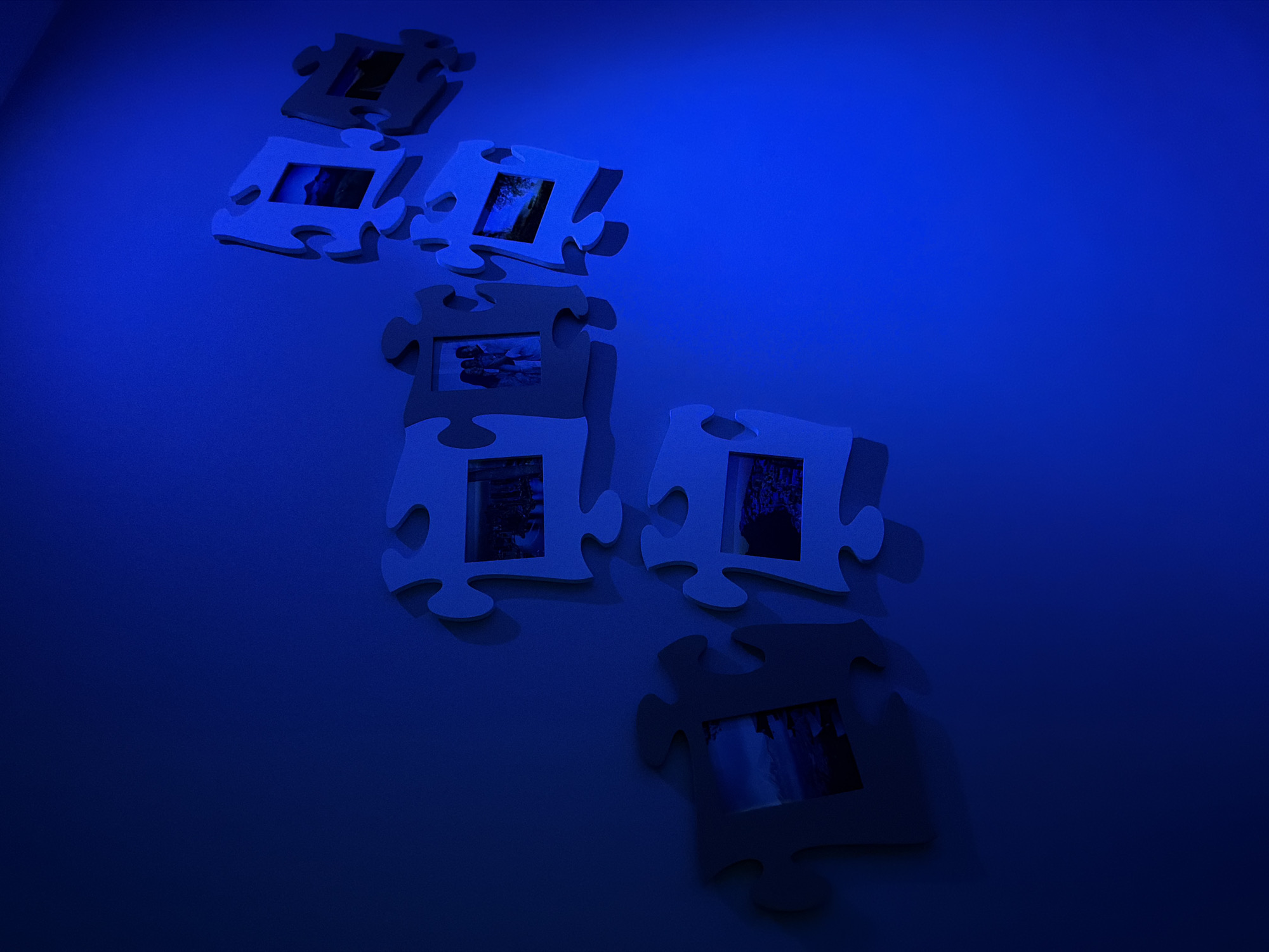The image shows a series of jigsaw puzzle pieces mounted on a wall, each containing a small photograph. The puzzle pieces are arranged in a vertical, slightly diagonal line. There are at least seven puzzle pieces visible. The lighting is predominantly blue, which casts a deep hue over the scene. The background is a solid blue wall. There are no people visible in the image. The location details for Pétange, Luxembourg are not represented in the scene.