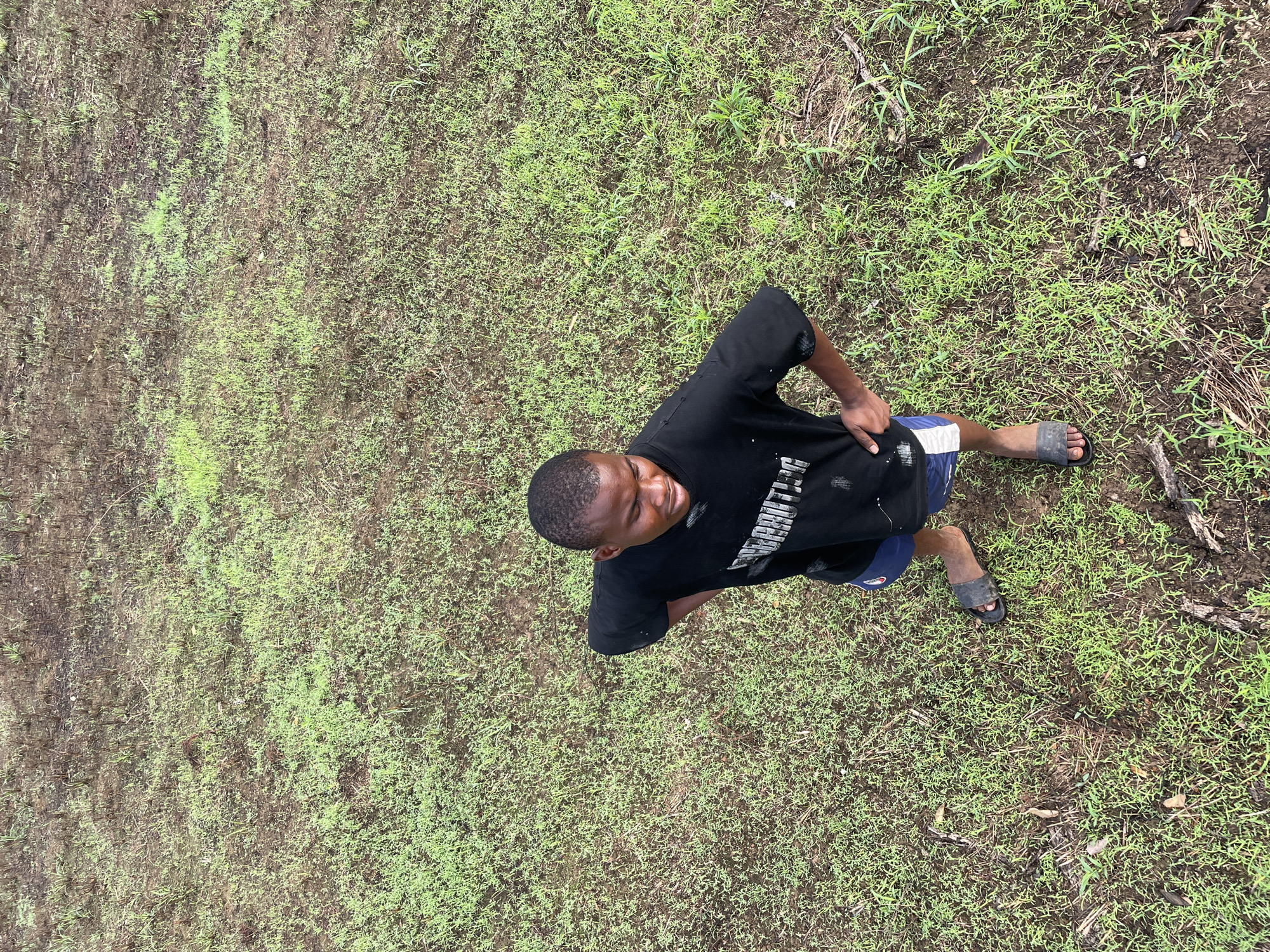 A young man stands in a grassy area, likely outdoors. He is viewed from above at an angle. The man is wearing a black t-shirt, blue shorts with a white stripe, and dark-colored sandals. The environment consists of varying shades of green grass with patches of bare earth. The location is Agba Umana, Nigeria.