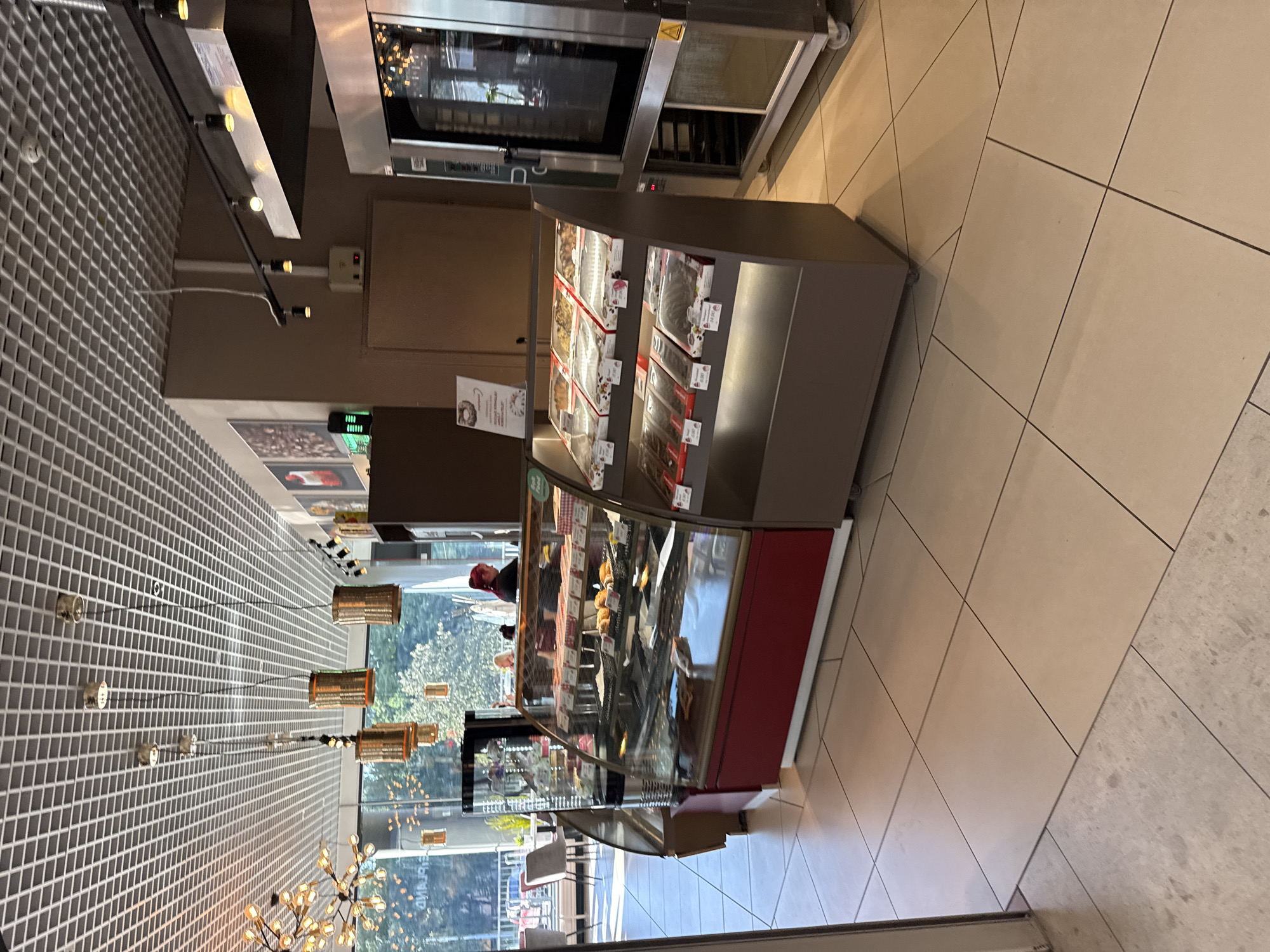 This image shows the interior of a cafe or bakery in Tallinn, Estonia. The scene is well-lit with overhead fluorescent lighting and decorative pendant lamps. In the foreground, a refrigerated display case filled with pastries and desserts is prominently featured. The case has a red base and a glass front, showcasing various items arranged in boxes and baskets. To the right of the display case is a large stainless steel oven, likely a commercial convection oven. In the background, through large windows, an outdoor scene with greenery and what appears to be a street or sidewalk with people and vehicles is visible. Inside the cafe, there are tables and chairs, and a few people are present, including a woman with pink hair behind the counter. The floor is tiled with light-colored square tiles. The overall atmosphere suggests a modern and clean establishment.