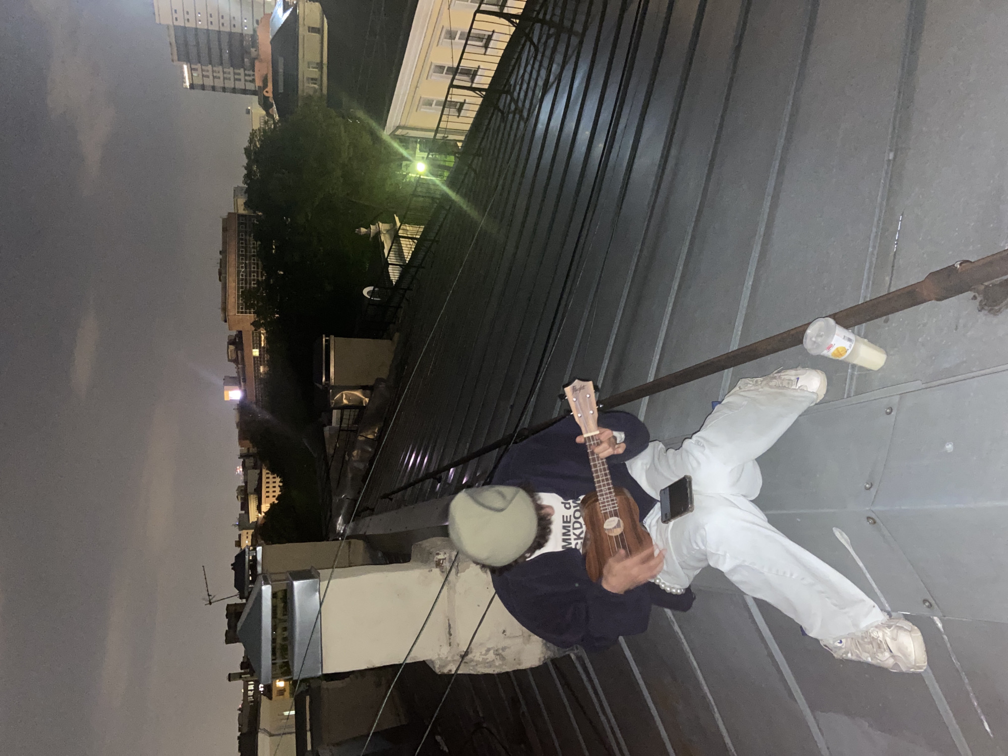 A person sits on a metallic rooftop in an urban environment, possibly in Moscow, Russia, based on the architectural style of surrounding buildings. The time of day appears to be evening or night, given the dim lighting and the presence of illuminated buildings in the distance. The person is wearing light-colored pants and sneakers, a dark jacket over a white top with writing, and a light-colored hat. They are holding a ukulele. A drink in a plastic cup is visible nearby. The individual is casually posed, occupying a large portion of the frame against the backdrop of the city.