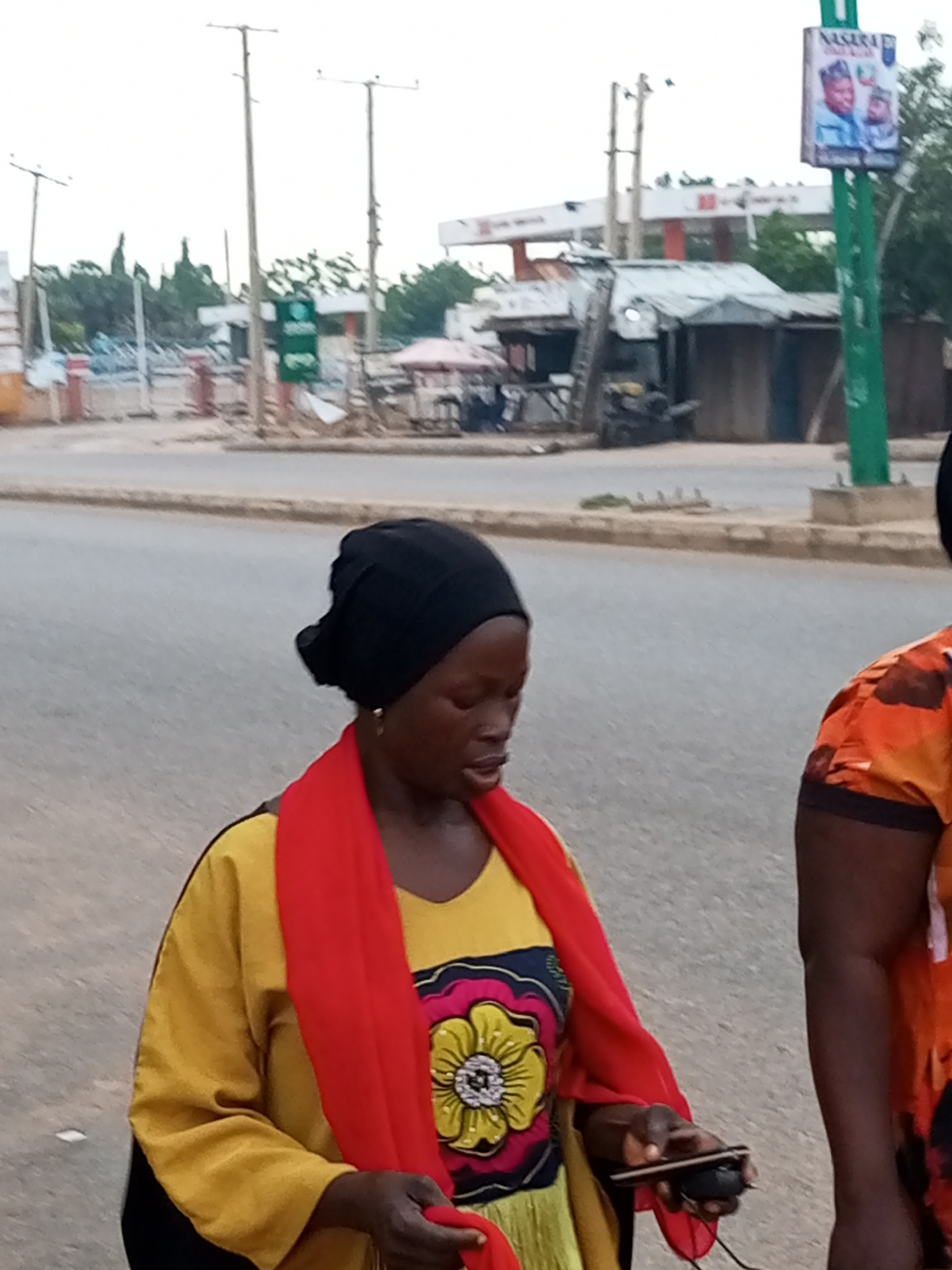 In Gombe, Nigeria, two women walk on a street. One woman, wearing a black headscarf, a yellow garment, and a red shawl, holds a mobile phone and a power bank. The other woman, partially visible on the right, wears an orange patterned shirt. In the background, a billboard with political candidates’ images and a green pole stand near a row of shops and a gas station. The street is paved and appears to be a commercial area.
