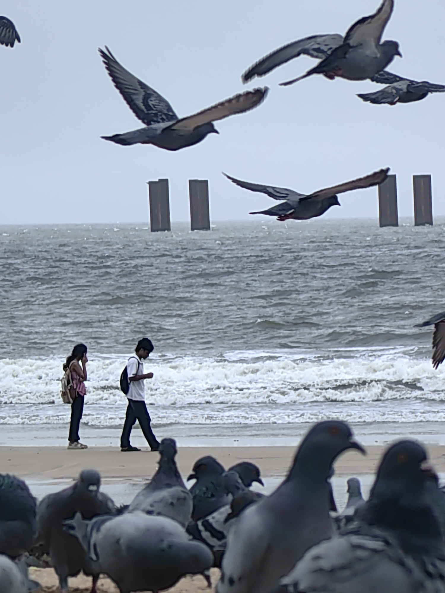 A group of pigeons are gathered on a sandy beach in Mumbai, India, with a body of water and a grey, overcast sky in the background. Two people, a woman and a man, are walking along the shoreline. The woman, wearing a pink top and carrying a backpack, appears to be talking on her phone. The man, dressed in a white shirt and dark pants and also carrying a backpack, walks ahead of her. Several pigeons are in flight above the water, and more are scattered on the sand in the foreground. Two large, dark vertical structures, possibly pylons or pillars, rise from the water behind the people. The ocean is rough with visible waves breaking on the shore.
