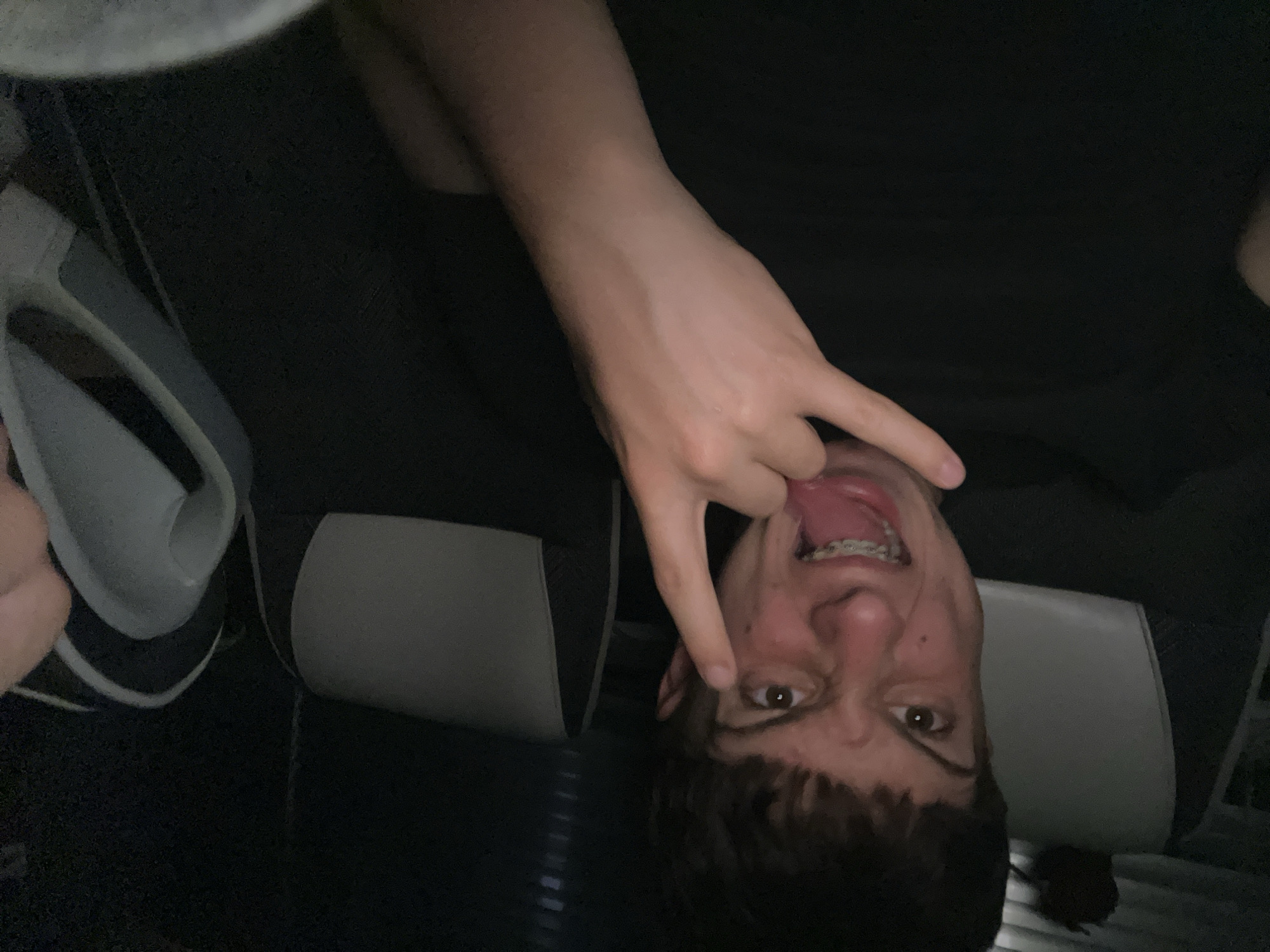 A male individual with short brown hair, brown eyes, and braces on his teeth is sticking out his tongue while making a peace sign near his eye. He is seated in a black chair with grey padding. A grey object with white lining is partially visible to the left. The setting appears to be indoors. The location is unknown and not specified in the prompt.