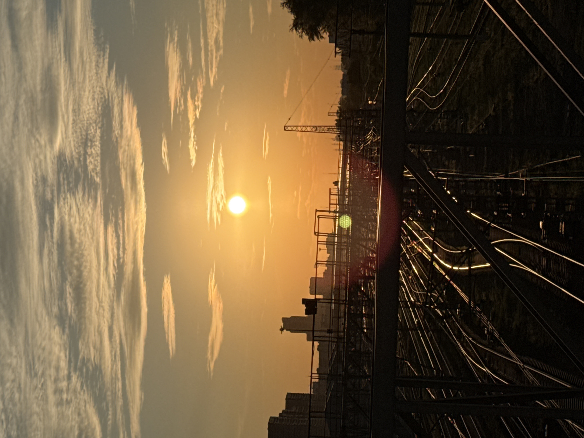 The image depicts a sunset over a train yard in Moscow, Russia. The sky is a warm orange and yellow, with wispy clouds. The sun is a bright, circular orb, low on the horizon. In the foreground, a complex network of train tracks stretches out, illuminated by the setting sun. A metal structure, possibly an overpass or bridge, spans across the top of the frame, casting shadows on the tracks below. In the middle ground, buildings of various heights form a cityscape, silhouetted against the sky. A tall crane is visible to the right of the buildings. The overall scene is one of urban industrial landscape bathed in the warm light of dusk.