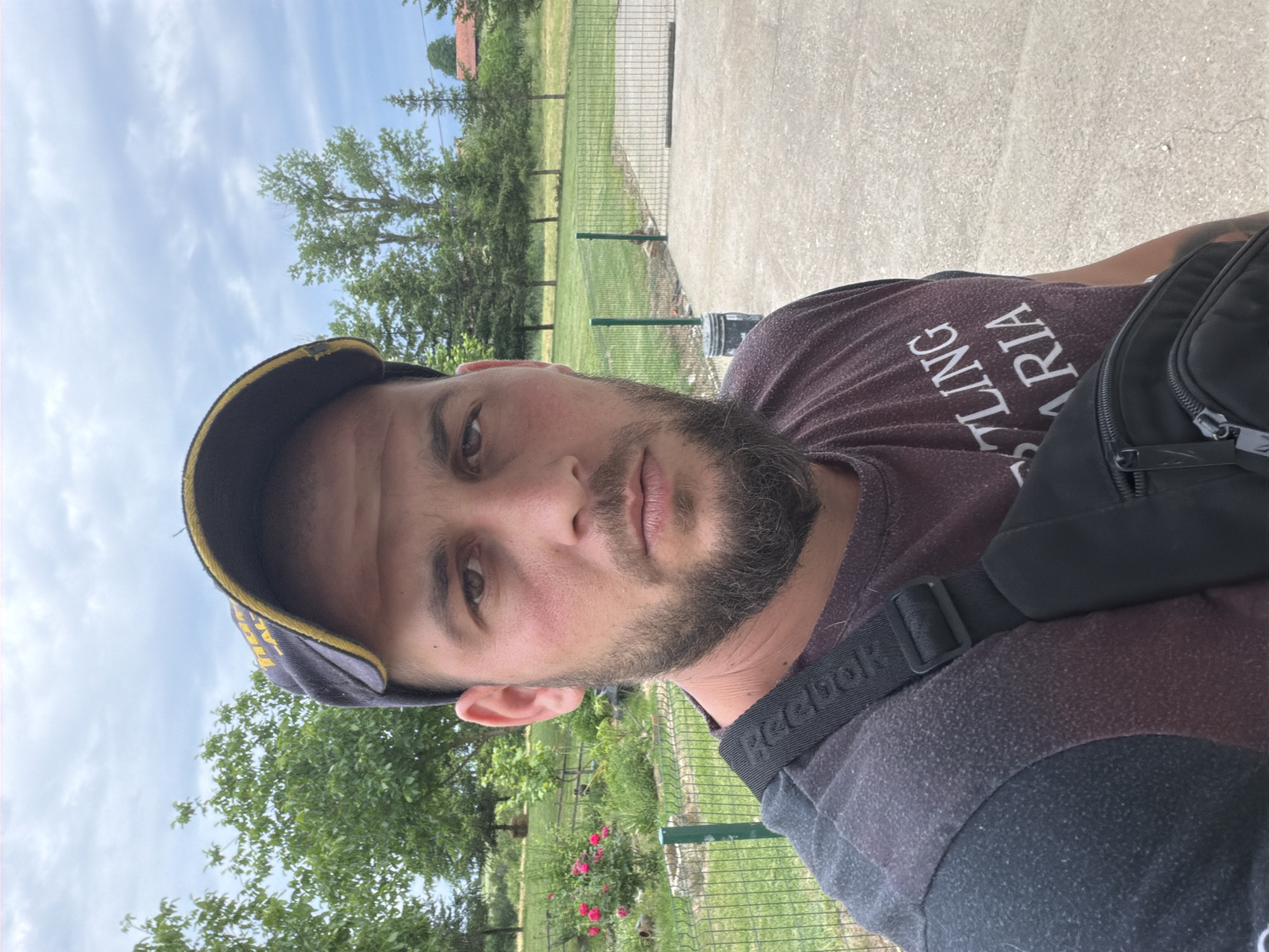 This is an outdoor, vertical close-up portrait, likely a selfie, of a man in Sirakovo, Bulgaria. The man, with a beard and dark hair, looks directly at the camera with a neutral expression. He wears a dark, possibly maroon or brown, short-sleeved t-shirt with white text. The visible text on the shirt is reversed and reads "STLING ARIA", suggesting it's part of a larger phrase like "WRESTLING BULGARIA". A black shoulder bag or fanny pack rests on his right side, with a black strap displaying the white "Reebok" logo. He also wears a dark blue or black baseball cap with yellow trim or text on the brim.
The setting is outdoors during daytime under a partly cloudy sky. A green wire mesh fence runs across the mid-ground, separating a concrete path in the foreground from a grassy area behind it. Numerous green trees are visible beyond the fence, and a bush with pink or red roses can be seen near the fence line on the left. In the distant background, through the trees, there is a glimpse of a building with a reddish roof. The scene suggests a park, a recreational area, or a semi-rural environment.