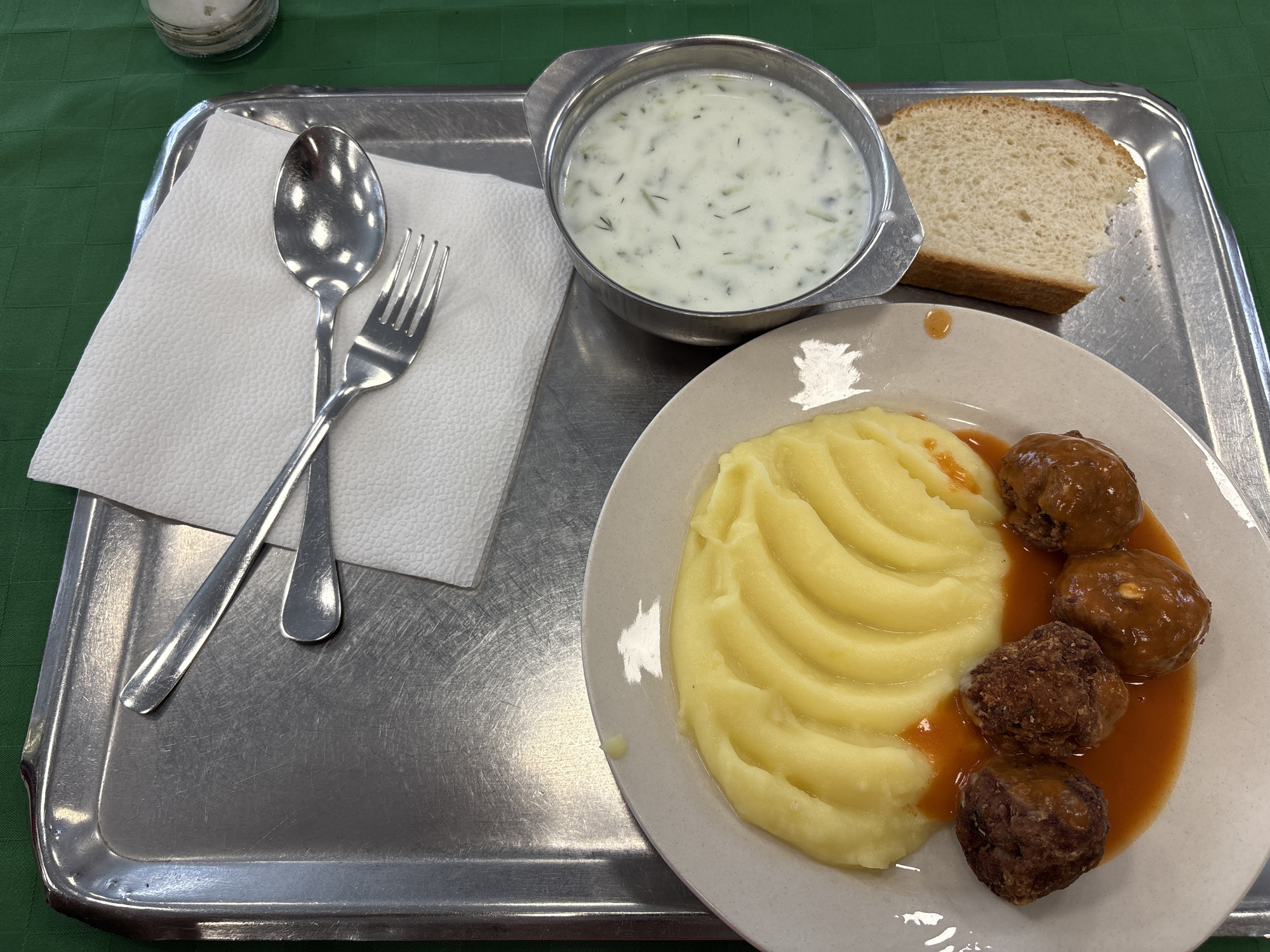 A meal on a metal tray sits on a green surface. The meal includes a white plate with mashed potatoes and four meatballs in a sauce. A metal bowl containing a white soup with green flecks rests beside the plate, next to a slice of white bread. A white paper napkin with a fork and spoon is positioned on the left side of the tray. A small container sits in the upper left corner of the tray. The location is Mirkovo, Bulgaria.