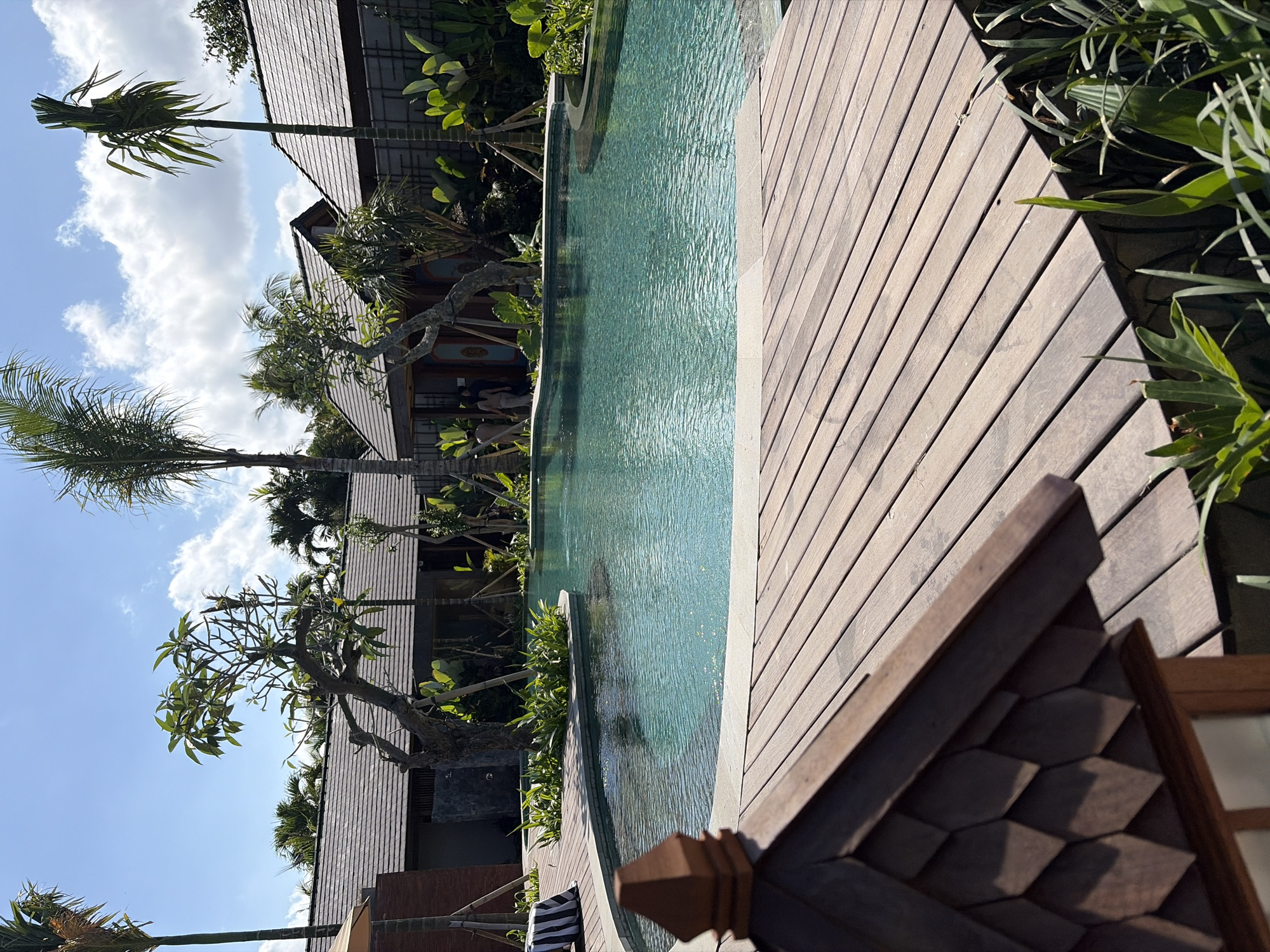 The image depicts an outdoor swimming pool area in Ubud District, Indonesia. The pool, filled with clear turquoise water, is surrounded by a wooden deck in the foreground and a stone-tiled edge. Several lush green plants and trees, including palm trees, are visible around the pool and between buildings. The buildings in the background have dark tiled roofs and wooden accents. A small wooden structure with a distinctive tiered roof is in the immediate foreground, partially obscuring the view of the pool. The sky is blue with scattered white clouds. Two individuals, appearing to be guests or staff, are walking near one of the buildings in the mid-ground. The overall scene suggests a resort or private villa setting.