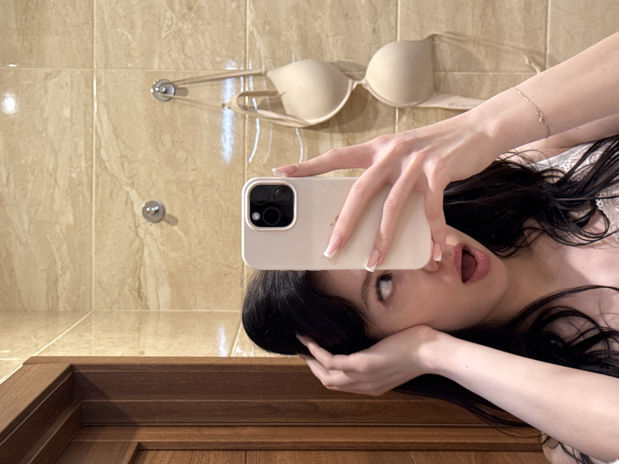 A young woman is taking a mirror selfie in a bathroom. She has dark hair and is wearing a white top. She is holding a smartphone in her right hand, which is angled to capture her reflection. Her left hand is touching her head, and her mouth is open in a surprised expression. In the background, a beige bra is hanging on a hook on the tiled wall. A wooden door frame is visible on the left side of the image. The bathroom appears to be modern with beige marble tiles. The photo was taken in Khimki, Russia.