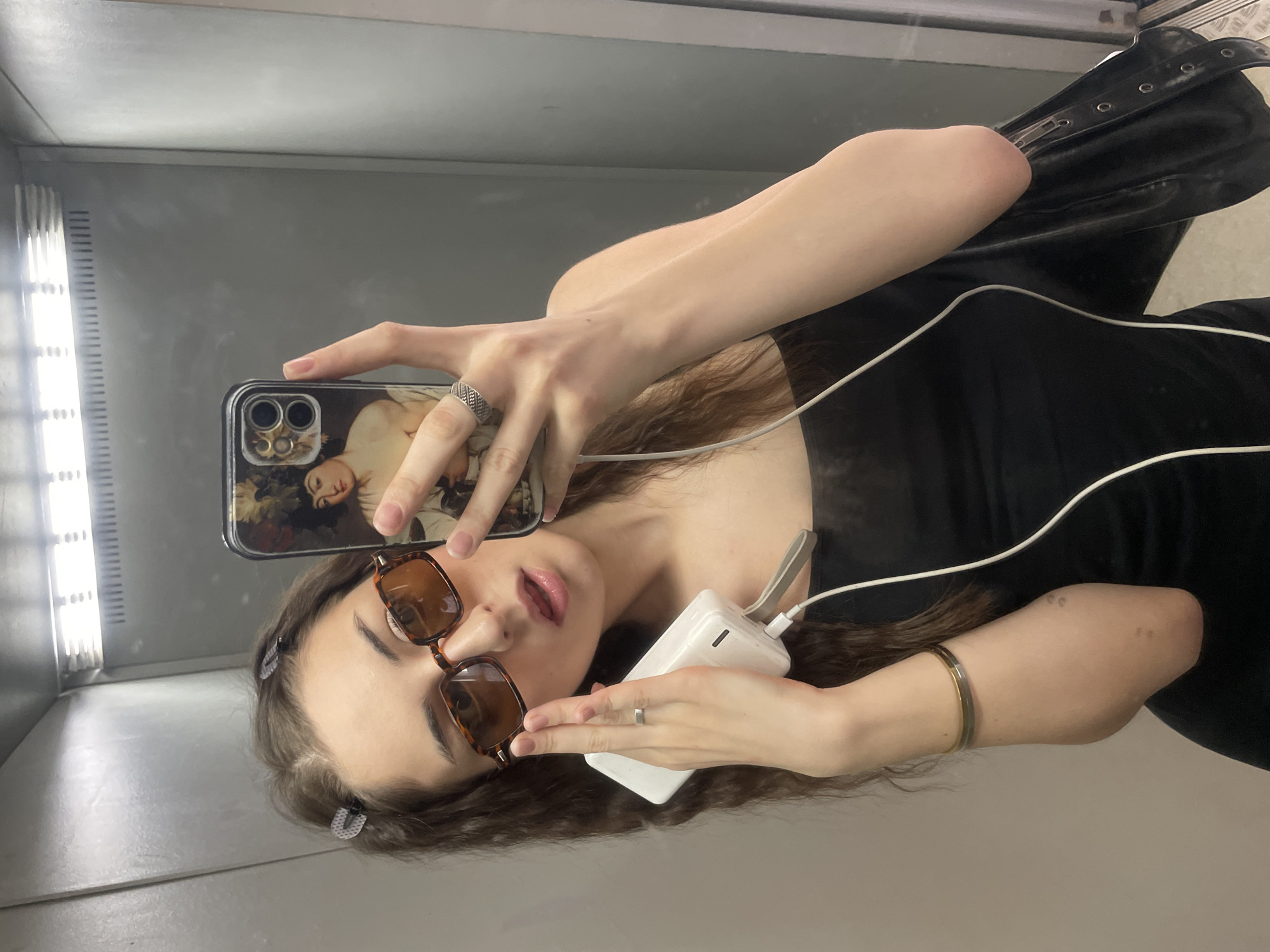 A young woman is taking a selfie in a mirrored space, possibly an elevator. She's holding a phone with a Renaissance art-themed case in her right hand, and a white portable charger connected to the phone with a white cable in her left hand. She's wearing sunglasses and a black top. The background consists of a gray wall with a vertical light fixture. There is no evidence to suggest that this image was taken in Moscow, Russia.