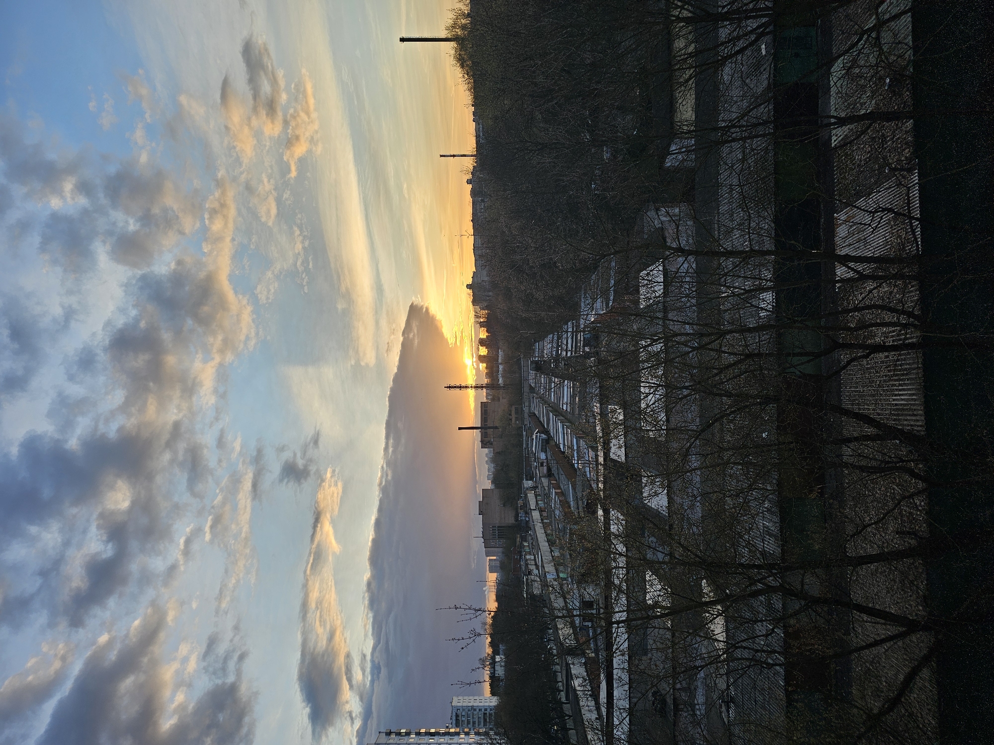 The image shows a view of the city of Khimki, Russia, during a sunset or sunrise. The sky features a mix of blue hues and clouds, some tinted with orange and yellow from the sun. Silhouettes of tall industrial chimneys are visible on the horizon. The foreground includes apartment buildings and rows of garages. Trees with bare branches are interspersed throughout the urban landscape.