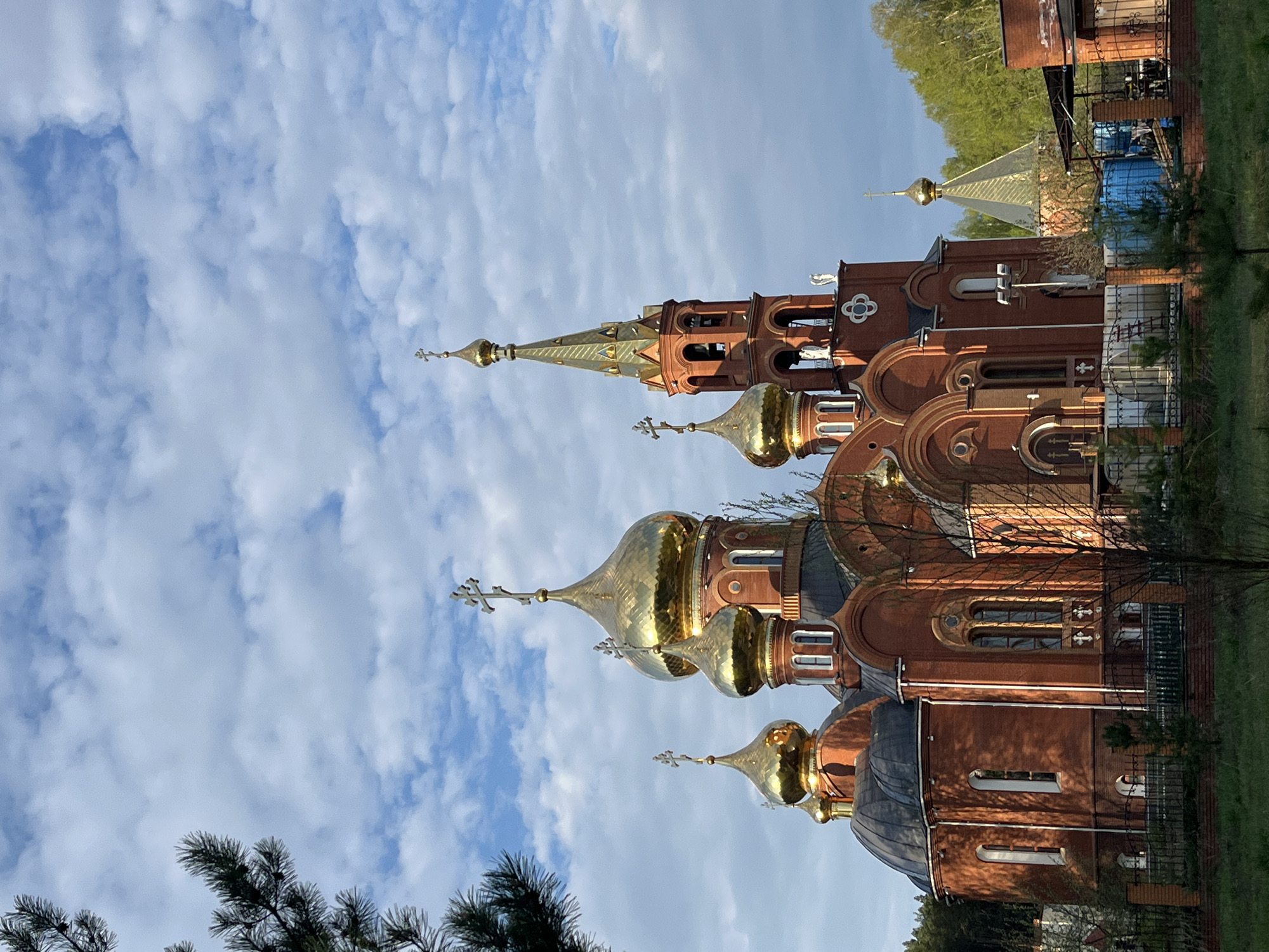 The image features a Russian Orthodox church, likely located in Chaikovsky, Russia. The church is constructed of red brick and adorned with multiple golden domes, each topped with a cross. The architecture includes arched windows and decorative elements. The church is positioned against a backdrop of a blue sky filled with puffy white clouds. Some trees and foliage are visible around the base of the church, suggesting it is set in a somewhat green area. There is also a glimpse of a structure behind the church.