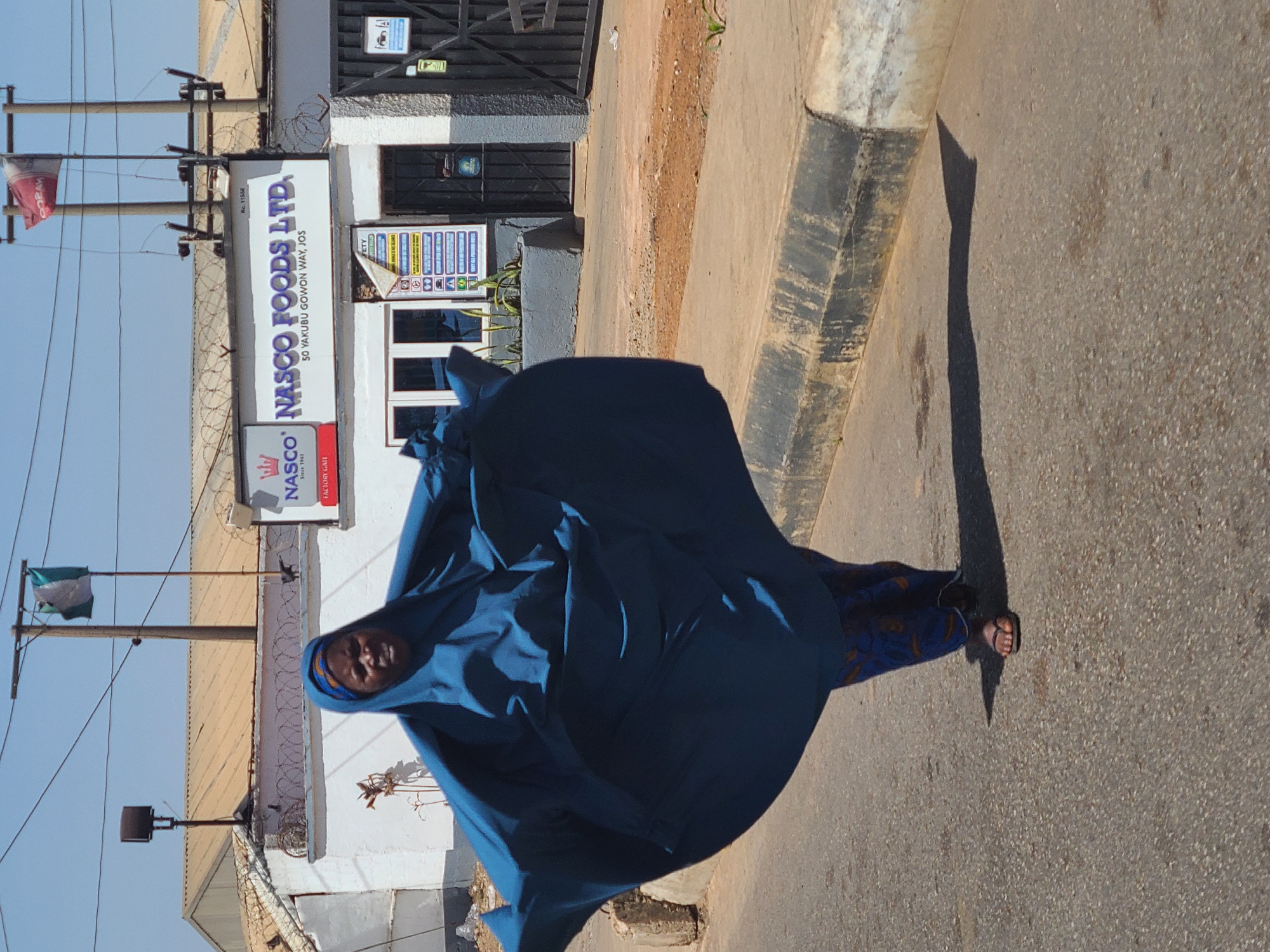 The image captures an outdoor street scene in Jos, Nigeria, featuring a person walking along a paved path next to a building. The individual, likely a woman, is dressed in a flowing, deep blue garment that covers most of her body, revealing patterned blue trousers and black flip-flops. Her face is partially visible, showing a neutral expression as she moves.
In the background, a white building houses "NASCO FOODS LTD.," as indicated by prominent signage. The sign also displays the address "50 YAKUBU GOWON WAY, JOS," confirming the location context. Below the main sign, a smaller red sign reads "FACTORY GATE," and above it, small text includes "W Rc. 11558." The building's exterior is secured with barbed wire along the top of a low wall. A safety information sign with various pictograms, partially reading "FETY" (likely "SAFETY"), is visible near the entrance.
Utility poles with wires extend across the frame. A green and white Nigerian flag is visible on one pole, and a red flag with unclear white text (possibly "DORAY") hangs from another. The sky is clear and bright blue, suggesting sunny weather. Long, distinct shadows on the ground, including one cast by the walking person, indicate strong sunlight, likely in the morning or late afternoon.