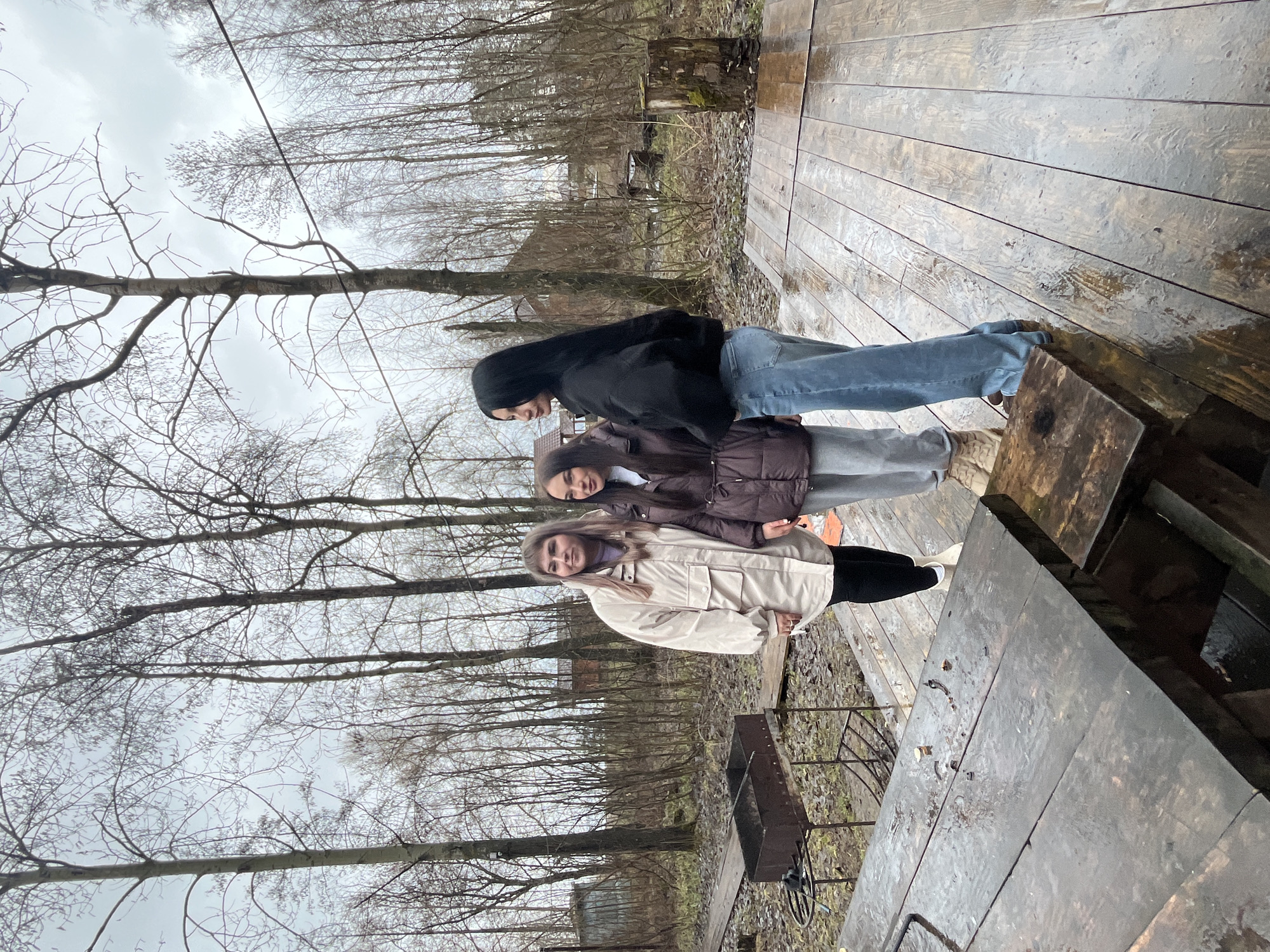 Three young women stand together on a wooden deck in what appears to be an outdoor recreational area in Chaikovsky, Russia, given the local flora and potential architectural hints in the background. The weather is overcast and possibly rainy, indicated by the damp surface of the wooden deck.
The woman closest to the camera is wearing a light-colored jacket with dark leggings and white shoes. The woman in the middle has a dark brown puffer jacket, gray pants, and light-colored footwear. The woman furthest back is wearing a black hooded jacket and blue jeans.
The background features bare trees typical of late autumn or early spring. A barbeque grill is situated on the ground near the deck. Other wooden structures are visible in the distance.