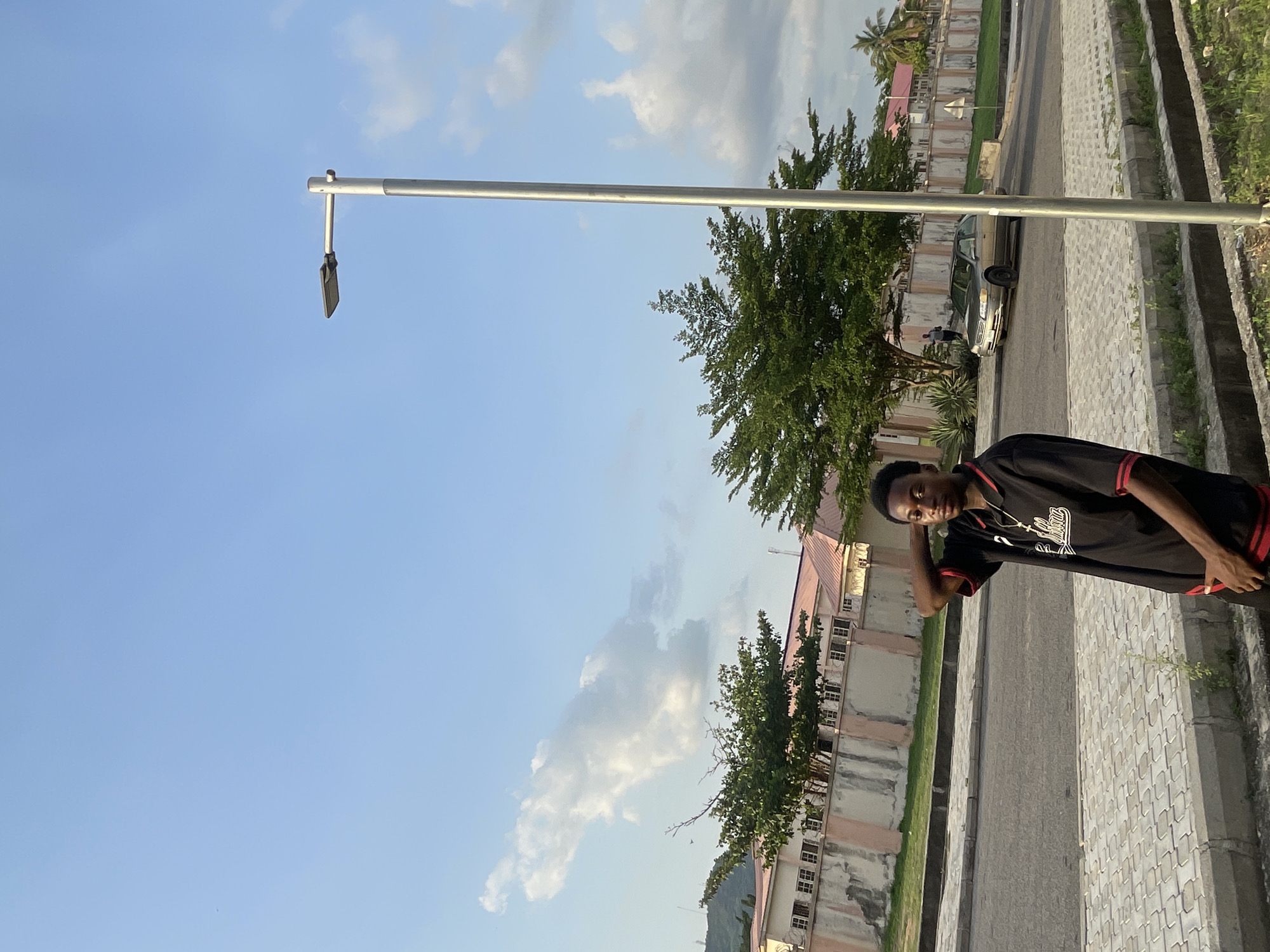 A medium shot depicts a young man standing on a paved walkway in Lokoja, Nigeria. The walkway is bordered by a curb on one side and a building with multiple windows and trees on the other. A light pole extends horizontally across the upper portion of the frame. In the background, a street with a parked car is visible. The sky is blue with scattered clouds. The young man is wearing a black shirt with red trim. He has one hand on his head.