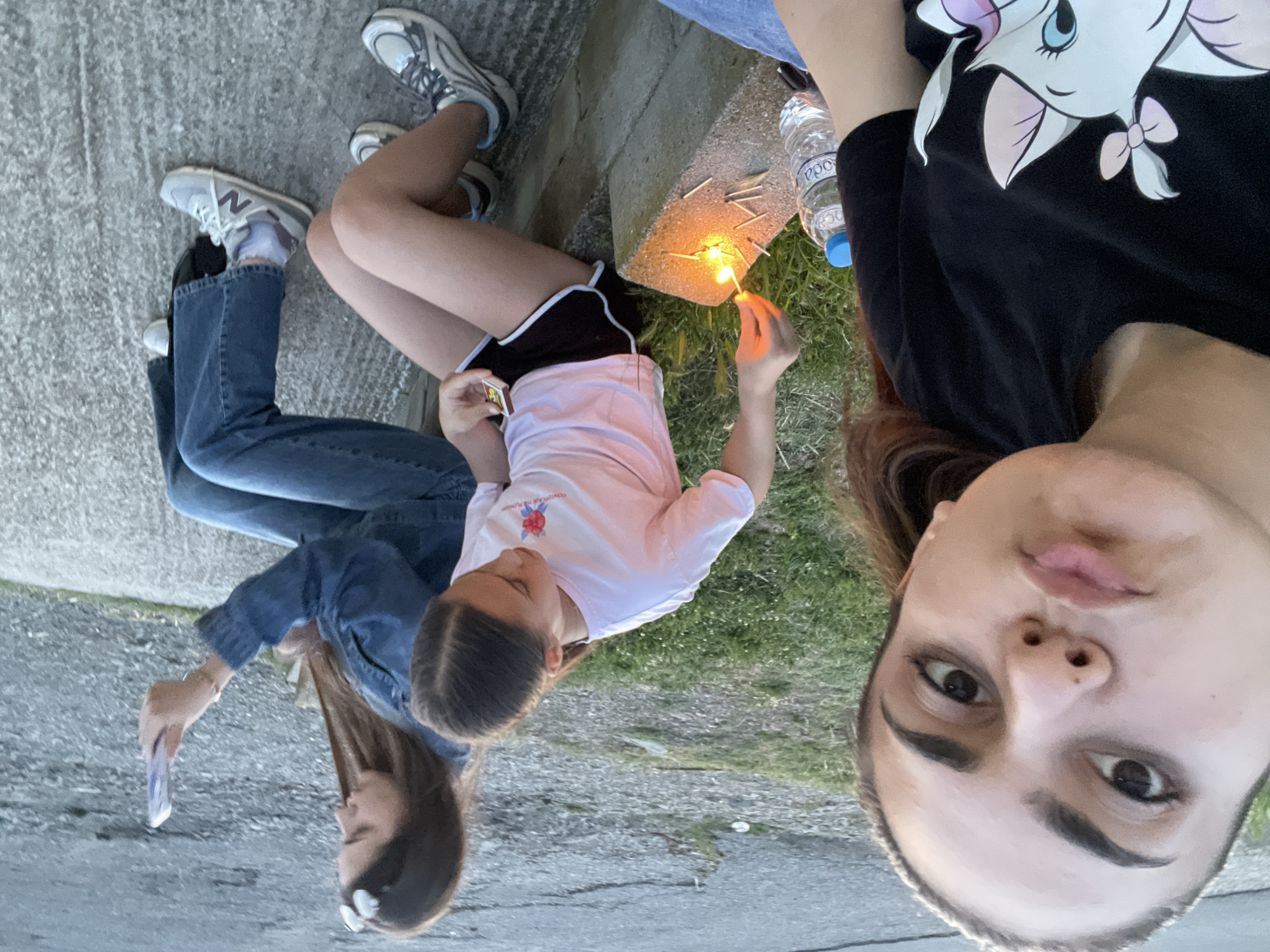 The image displays three females situated outdoors in Debelt, Bulgaria. In the immediate foreground, a young adult female with dark hair, pulled back, is positioned to take a selfie. She is wearing a black t-shirt featuring a white cartoon cat design with a pink bow and has her lips pursed. To her left, a clear plastic water bottle with a blue cap and "Boca" branding is visible.
In the middle ground, a younger female with dark hair, parted down the center, is seated on a grey concrete block. She is dressed in a light pink t-shirt with a red floral design and the partial text "CONTEMPLATING THE FLOWERS," black shorts with white trim, and grey and white athletic shoes. Her right hand holds a lit matchstick, which she is using to ignite a collection of other matchsticks scattered on the concrete surface, some already burning. Her left hand holds a matchbox.
Further to the right, a second young adult female is seated on the same concrete block. She has long brown hair, wears a denim jacket, blue jeans, white socks, and grey and navy athletic shoes with a prominent 'N' logo. White accessories, possibly sunglasses or hair clips, are positioned on top of her head. She holds a light-colored smartphone vertically in her right hand, viewing its screen.
The ground surrounding the concrete block comprises green grass and visible dirt. The background beyond the individuals shows an out-of-focus expanse of grey asphalt or gravel with minimal vegetation. The scene is illuminated by natural ambient daylight.
