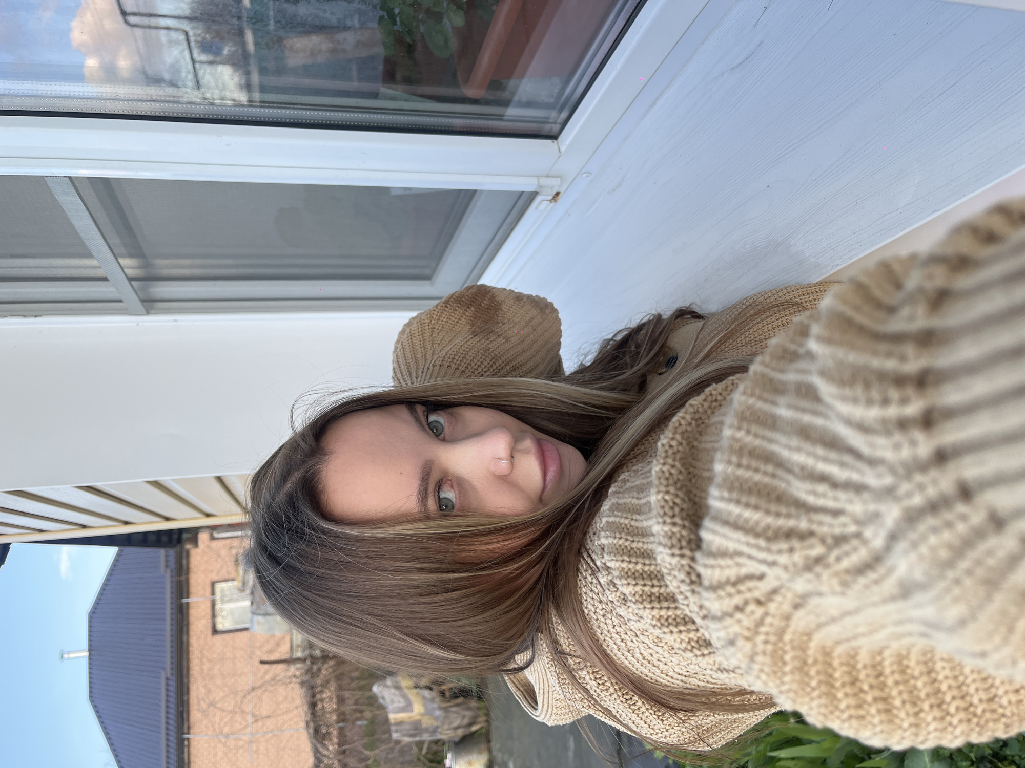 A selfie taken by a fair-skinned person with light brown hair and green eyes. She is wearing a beige knitted sweater and hat. The background shows a window with a white frame, reflecting a building with a blue roof and a brick facade. The outside scene includes greenery and a fence. The location is potentially Novopokrovskaya, Russia, based on the request context.