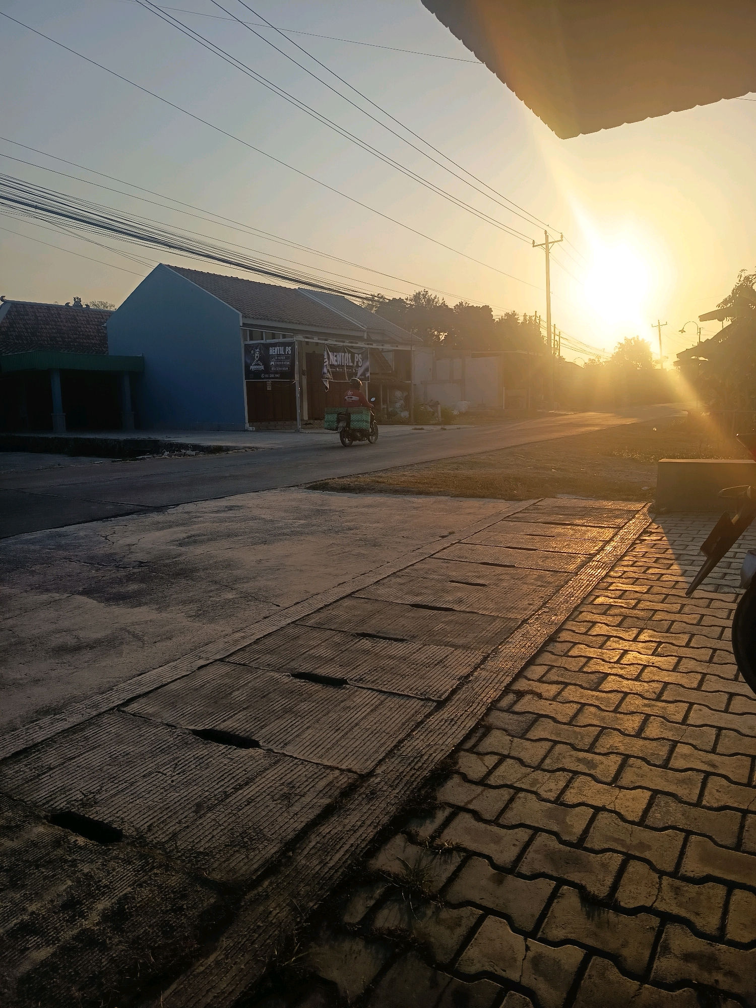 The image depicts a street scene in Kalisari, Indonesia, during sunrise. The sun is low in the sky, casting a warm, golden light and long shadows across the road and surrounding buildings. A blue building with a tiled roof is visible on the left, with signage reading "RENTAL PS". A person is riding a scooter, carrying a green crate on the back, and traveling away from the viewer on the asphalt road. The road is bordered by a concrete drainage grate and a paved walkway with interlocking bricks. Power lines are strung overhead, and a utility pole is visible. Trees and other residential structures are present in the background. The overall atmosphere is tranquil and typical of a residential area at the start of the day.