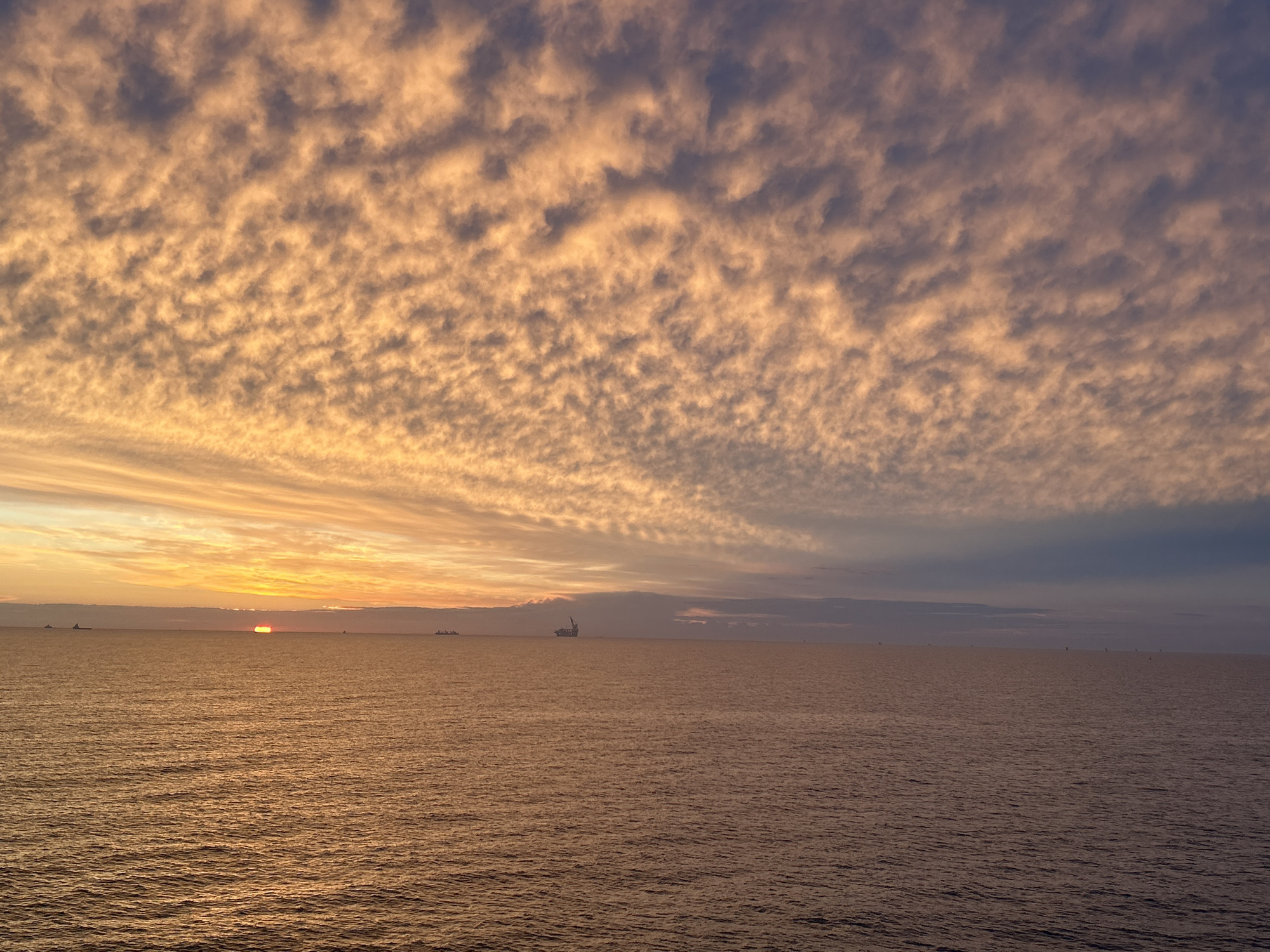 The image captures a panoramic seascape at either sunrise or sunset in the city of Unknown, Unknown. The sky, occupying the upper two-thirds of the frame, is filled with an extensive formation of altocumulus clouds, presenting a rippled or "mackerel sky" pattern. These clouds are predominantly illuminated in warm orange, golden, and peach tones, gradually transitioning to darker grey-blue in the upper right quadrant. The sun, a bright orange-red arc, is partially visible just above the horizon line, slightly to the left of the frame's center.
The lower third of the image features the vast, calm ocean surface, which reflects the warm colors of the sky, appearing in shades of dark orange, copper, and brown. Subtle ripples and surface texture are visible across the water. Several objects punctuate the distant horizon. To the far left, two dark, low-lying silhouettes, likely large vessels such as cargo ships or tankers, are visible. Near the center-right, a more prominent, complex dark silhouette, possibly an offshore oil rig or a specialized crane vessel, stands against the lighter sky. Further to the right, a series of very small, dark, regularly spaced structures, potentially buoys or offshore wind turbines, line the distant horizon. No people, specific actions, or interactions are discernible within the scene.