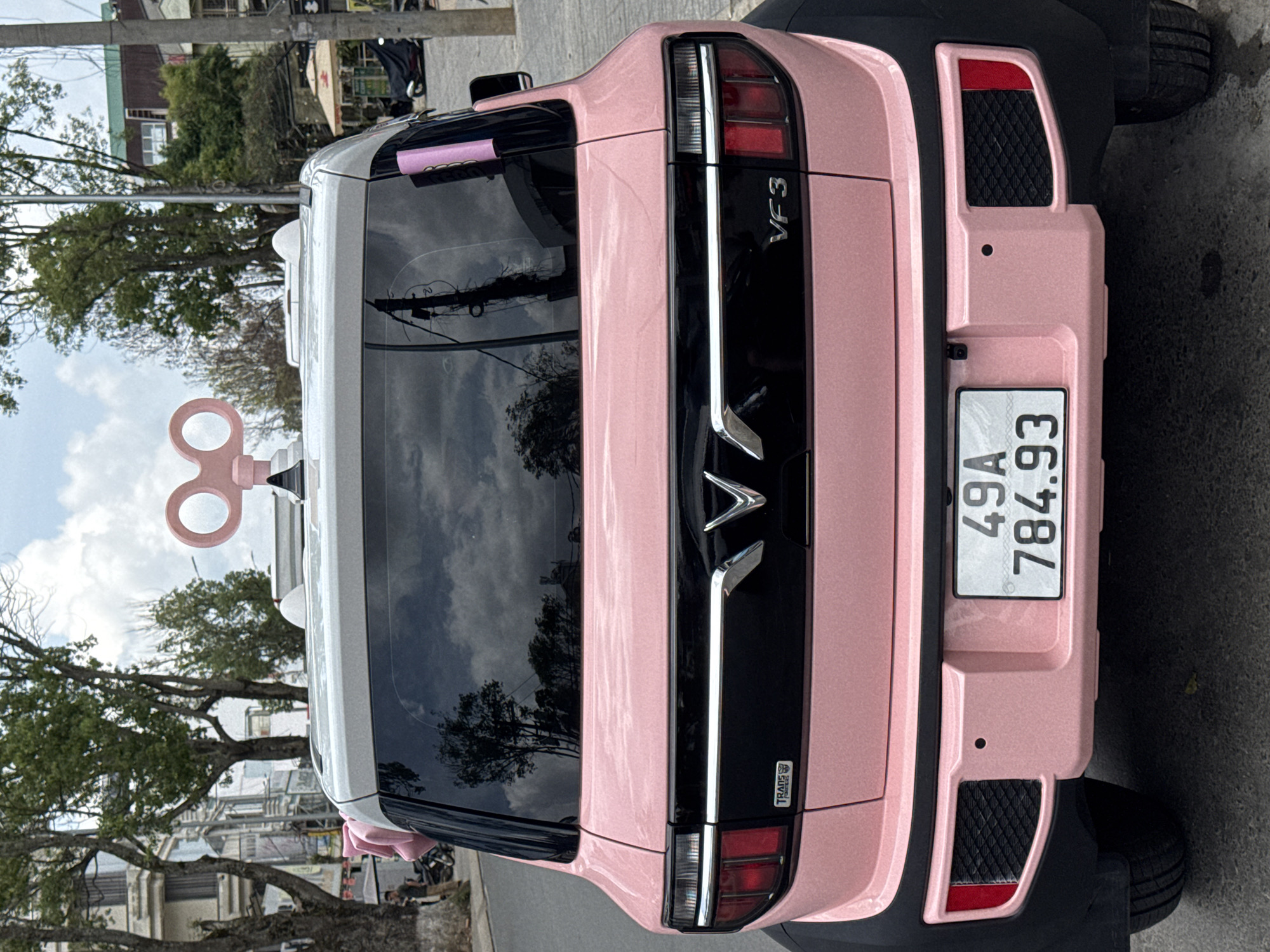 A rear view of a pink car with a wind-up key decoration on its roof is parked on a street. The car has a license plate that reads "49A 784.93". The background shows trees, buildings, and other cars, suggesting an urban environment. The street appears to be in Lâm Viên - Đà Lạt, Vietnam. The lighting indicates it is daytime, possibly overcast due to the reflections on the car's windows.