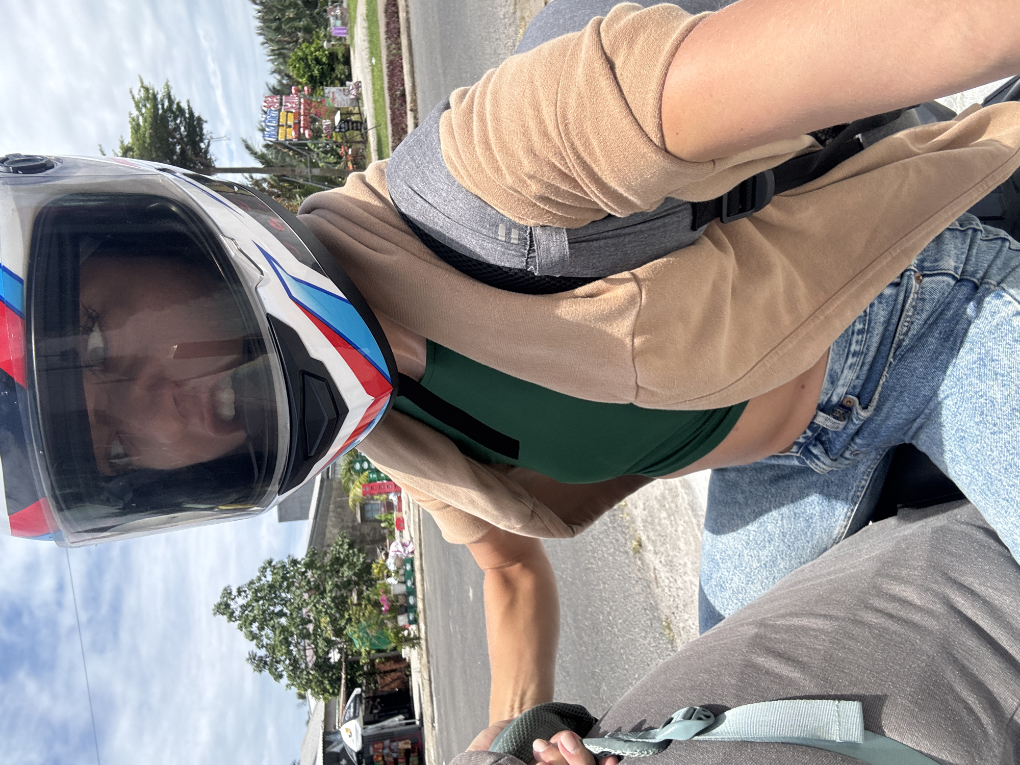 A person wearing a full-face motorcycle helmet is seen from a first-person perspective, likely while riding a motorcycle. The person is wearing a green top, a tan cardigan, and blue jeans. They also have a grey backpack on. The background shows a city street, trees, and a sign with colorful lettering, potentially indicating a local business. Based on the lettering on the sign and background foliage, this image is located in Đà Nẵng, Vietnam.